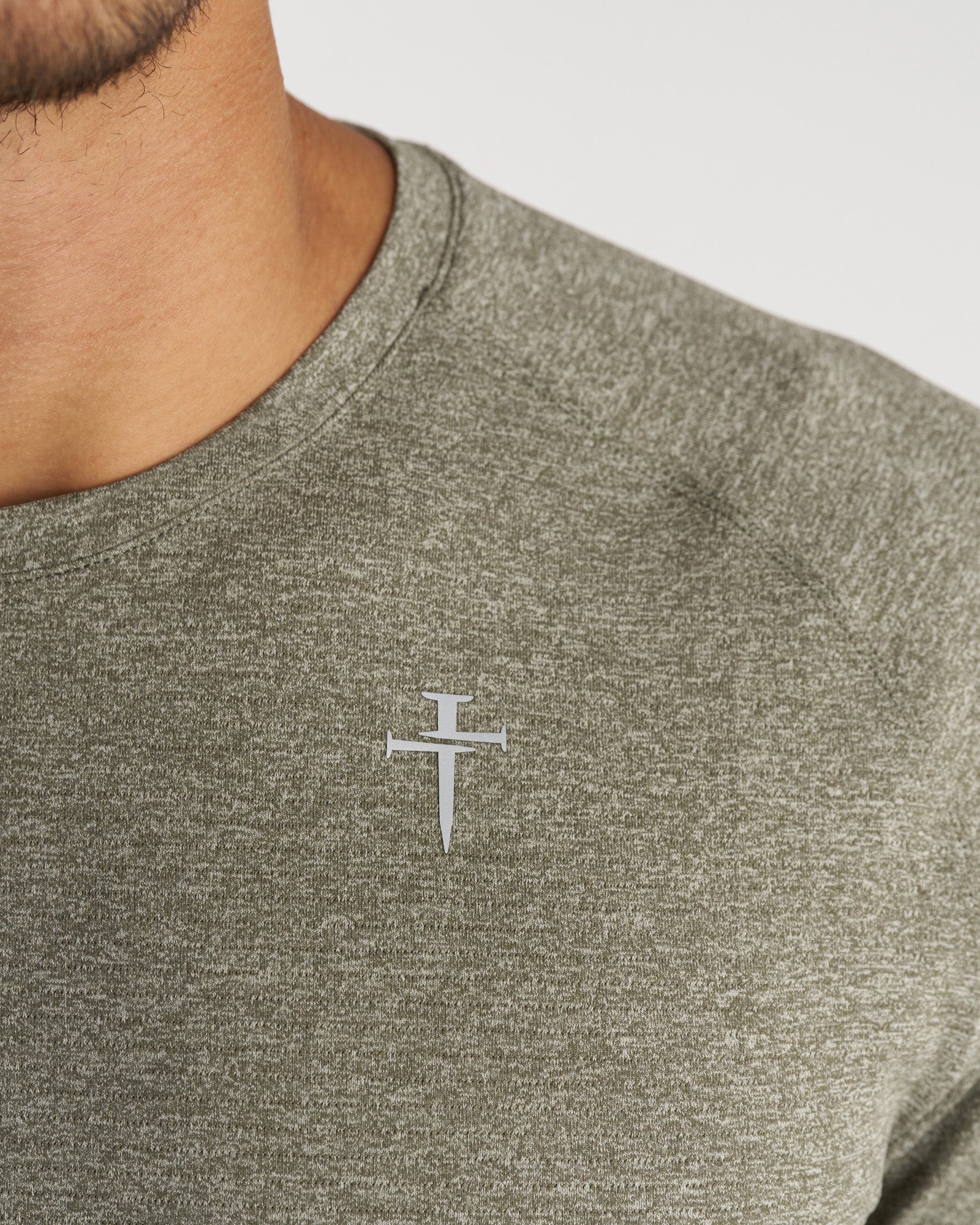 Close-up detail shot of the Aion-Lite Tee Cross in Sage Marl showing the small silver nail cross logo on the chest.
