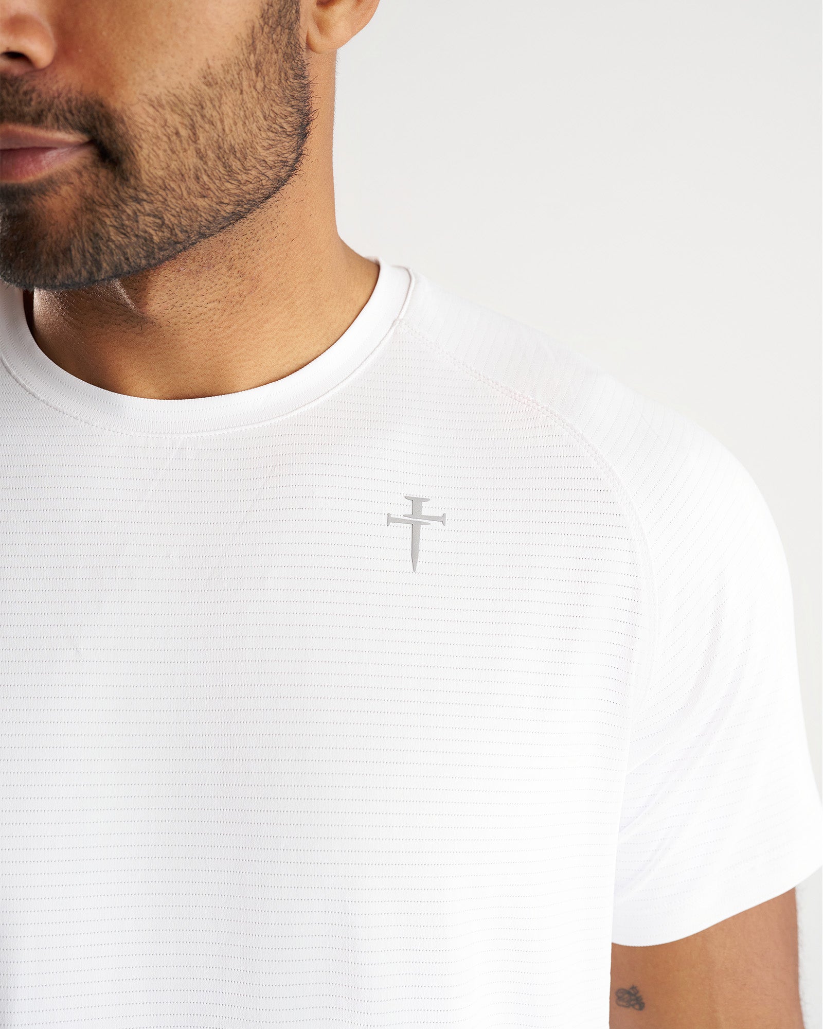 Close-up of Aion-Lite Tee Cross in white showing tonal cross logo on textured chest fabric.