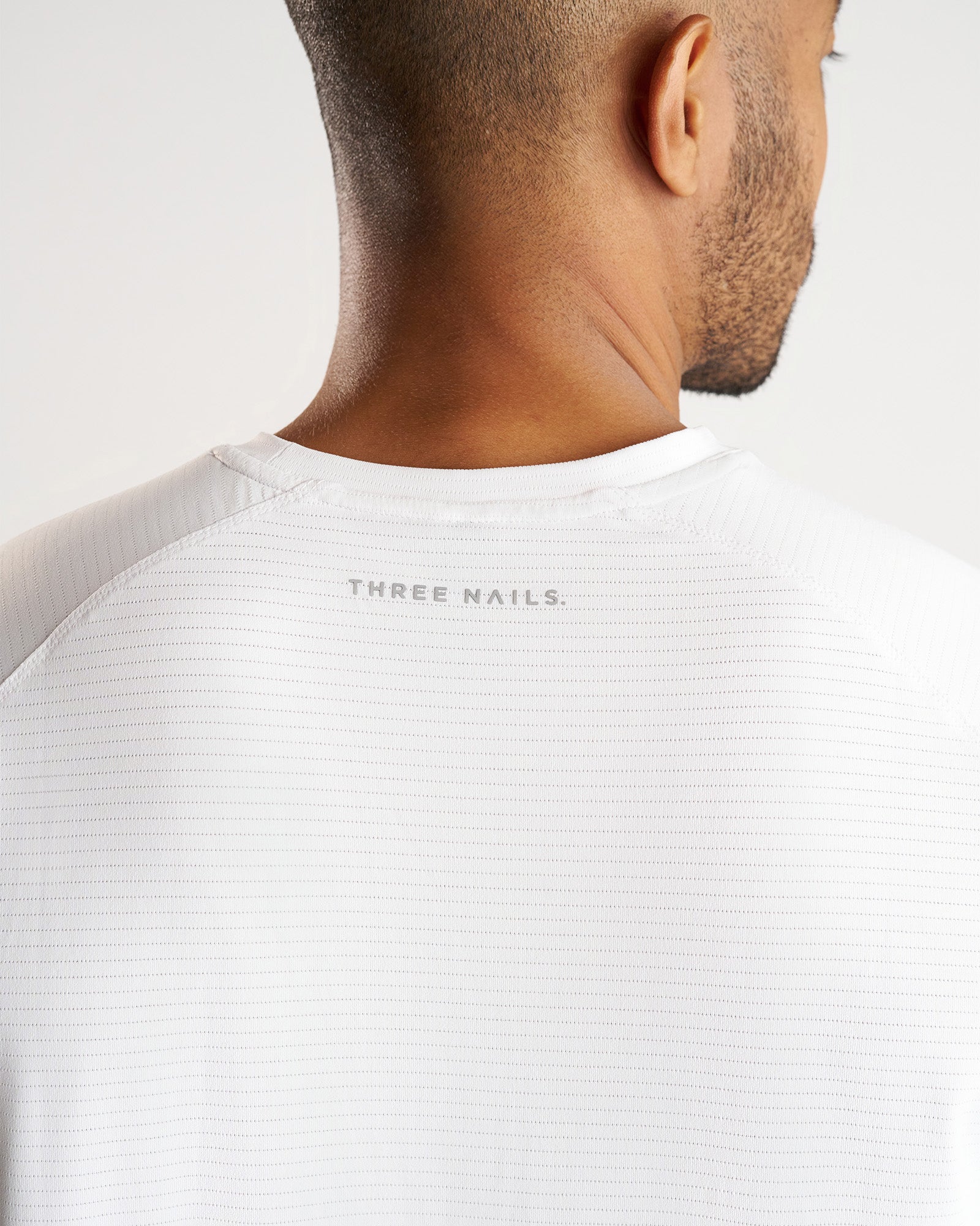 Close-up back neck detail of the Aion-Lite Tee Cross in white showing the Three Nails branded label and textured fabric.