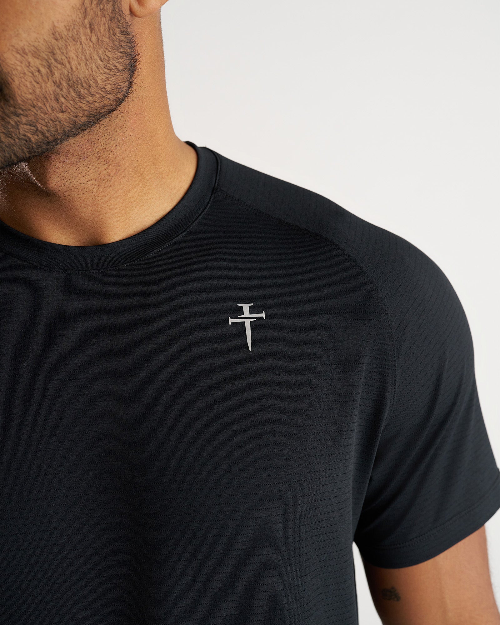 Close-up of the Aion-Lite Tee Cross in black showing the small silver cross logo on the chest.