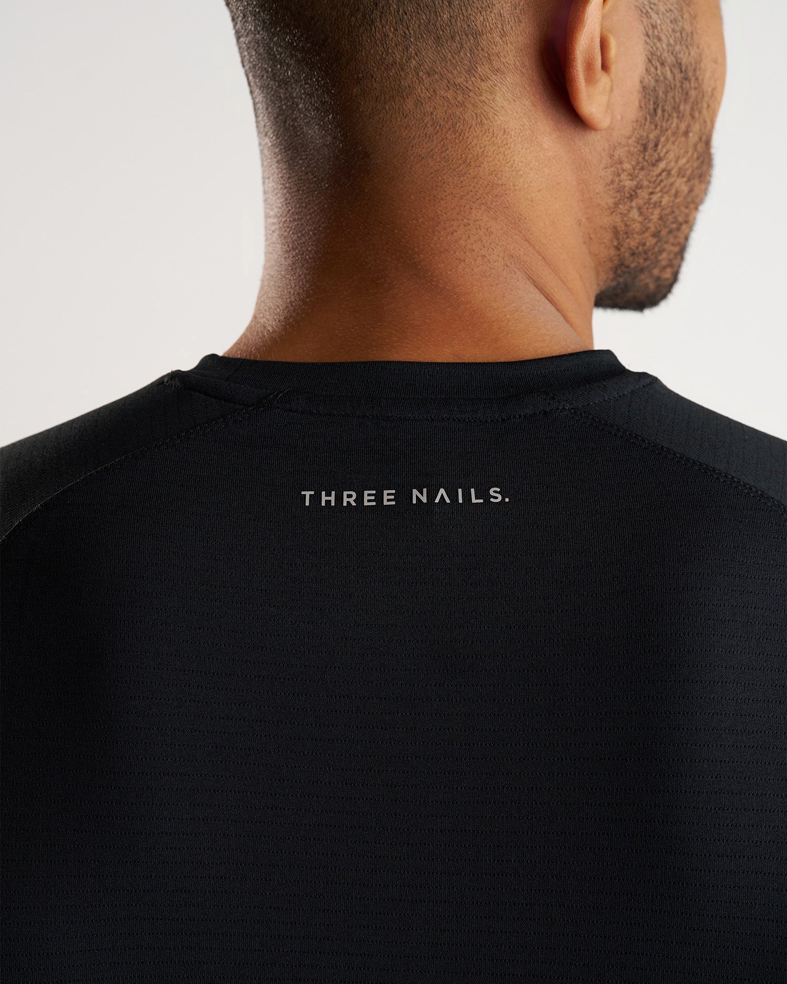Close-up back neck detail of the Aion-Lite Tee Cross in black showing the Three Nails. logo print.