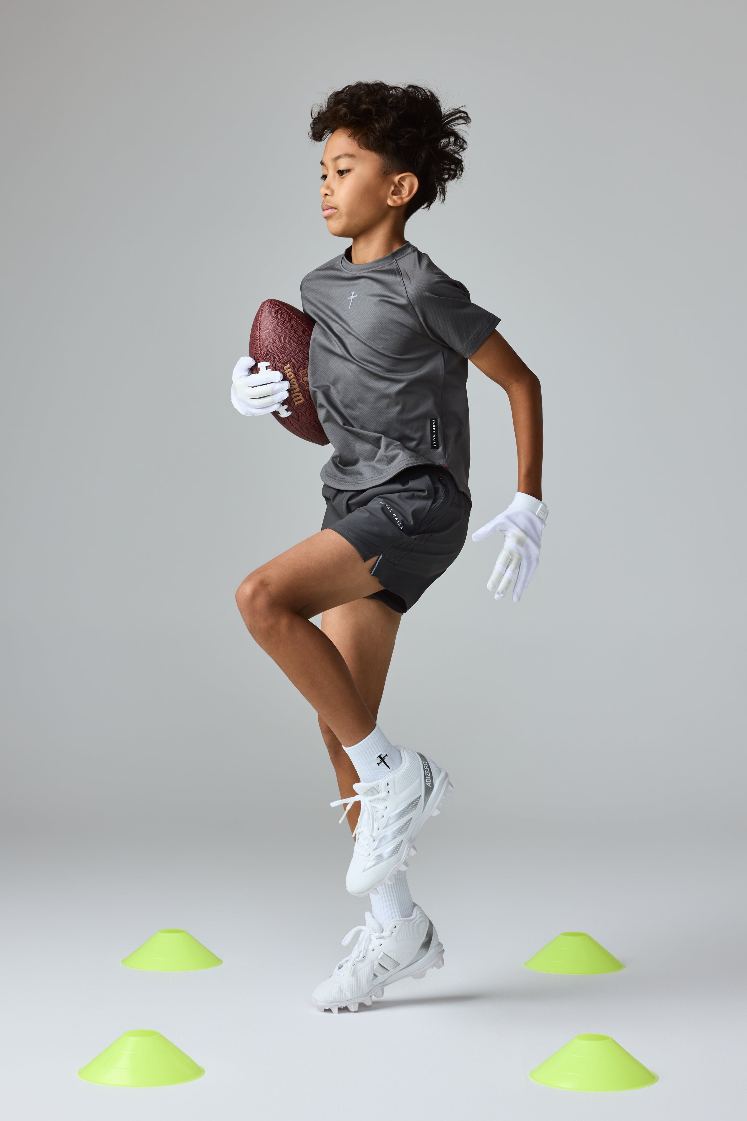 Youth Pro-Tech Tee Cross in Space Grey on a young boy in a dynamic running pose holding a football, front view.