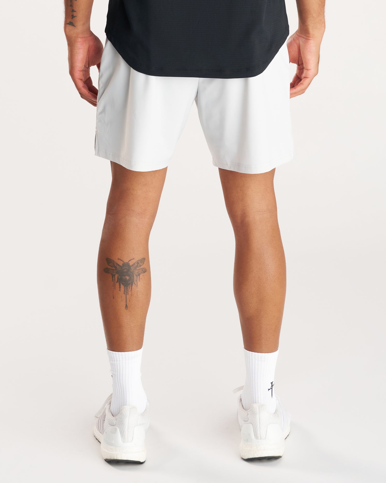 Back view of model wearing Vapor-Lite Liner Short Cross in Stone with white sneakers and a leg tattoo visible.