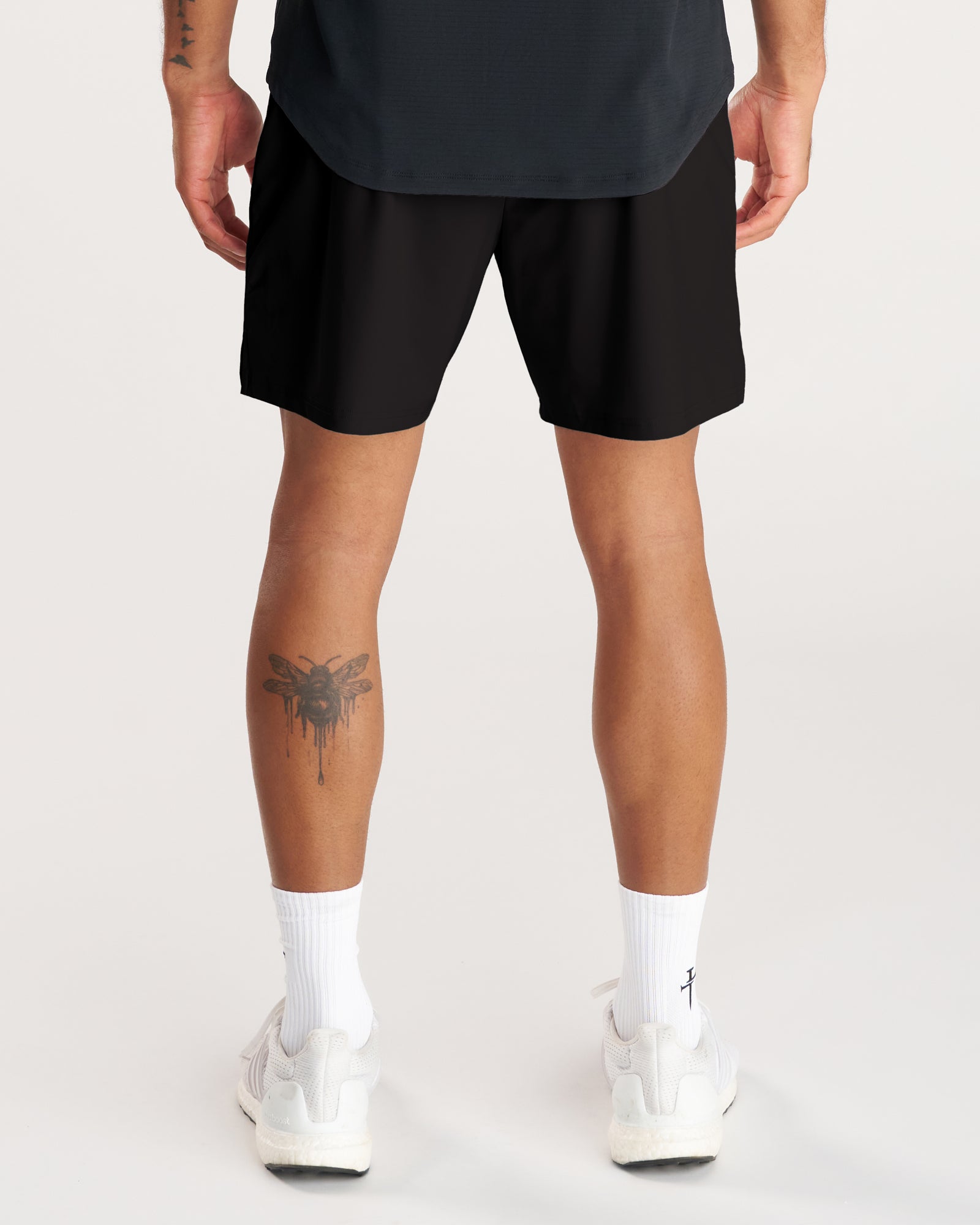 Back view of model wearing Vapor-Lite Liner Short Cross in black with white sneakers and a leg tattoo visible.