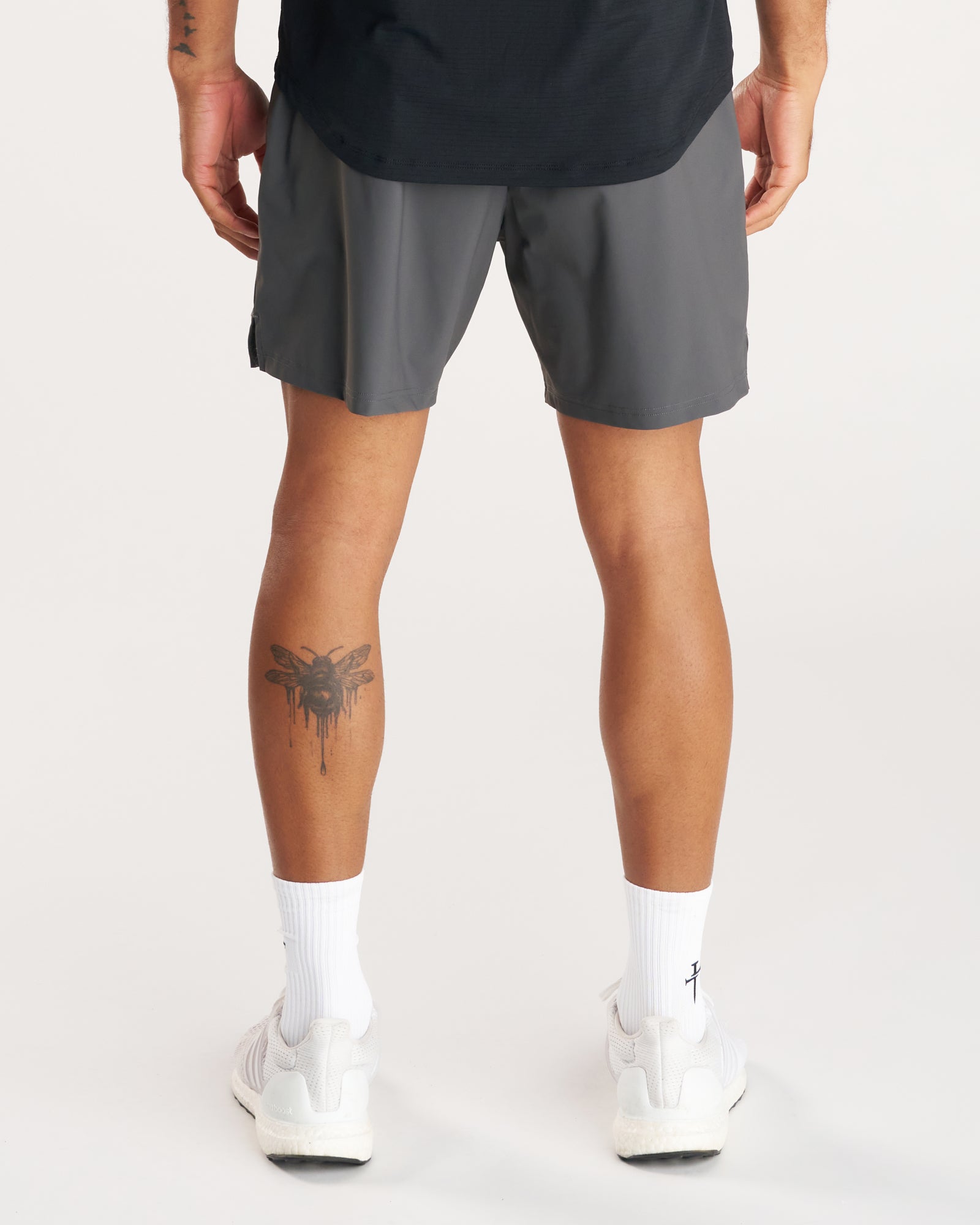 Back view of model wearing Vapor-Lite Liner Short Cross in Space Grey with white sneakers and a leg tattoo visible.