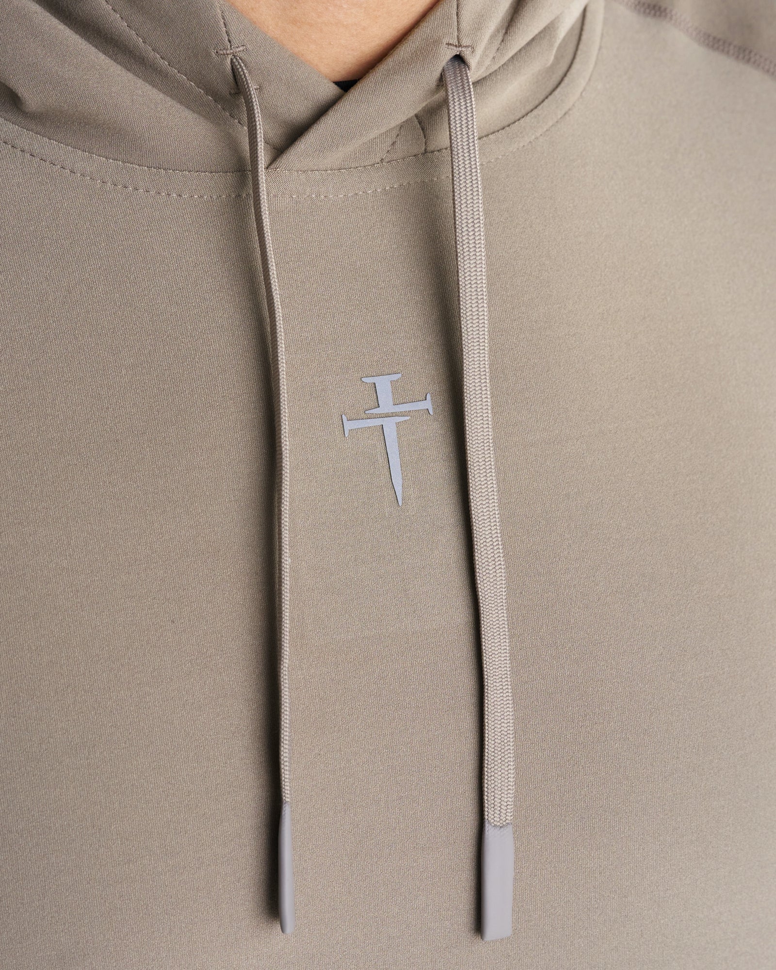 Close-up of Haven Performance Hoodie in Sand showing the Three Nails cross logo and drawstring detail.