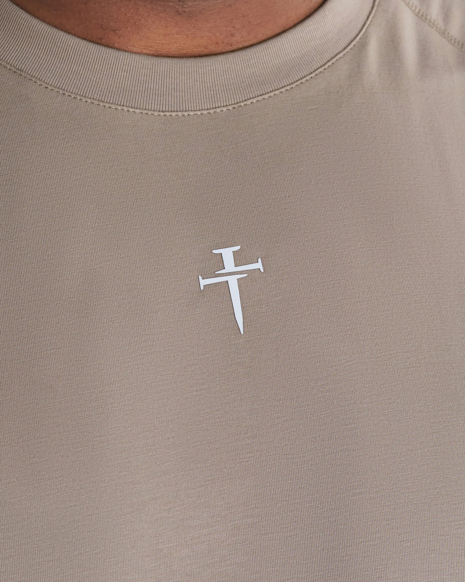Close-up of the white nail cross logo on a Core Raglan Cutoff Sand tank top, chest area front view.