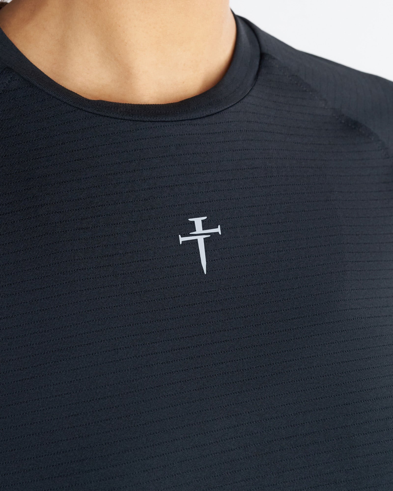 Close-up detail of Women's Aion-Lite Tee Cross in Black showing ribbed texture and small nail cross logo on chest.
