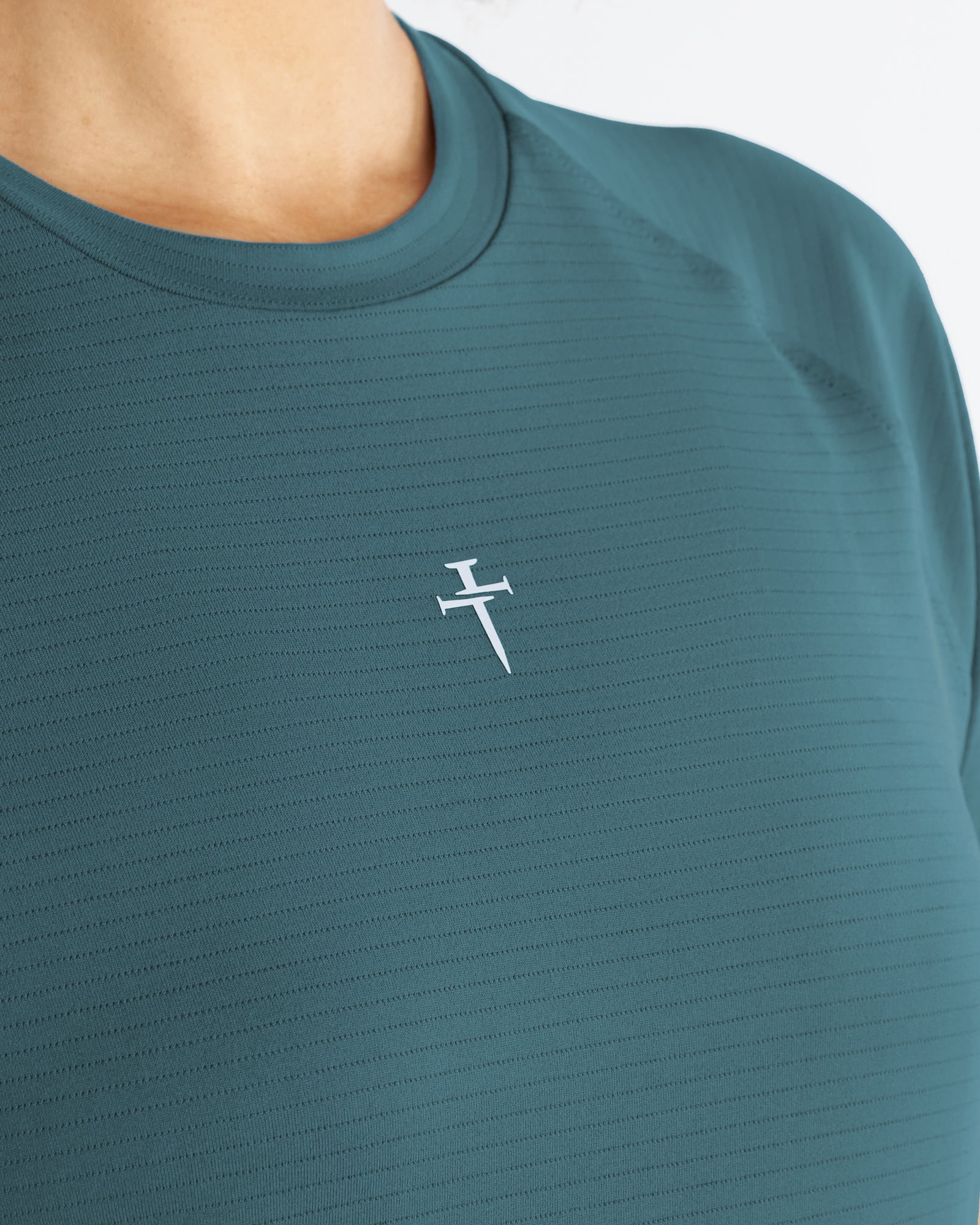 Close-up of the teal Womens Aion-Lite Tee Cross showing the white cross logo on textured fabric.
