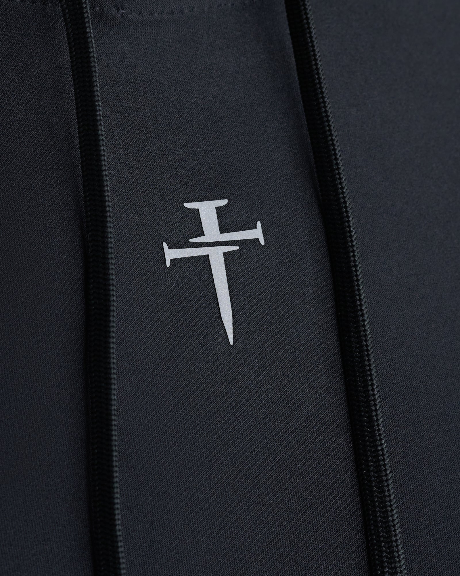 Close-up of the Haven Performance Hoodie in black showing a reflective nail cross logo on the chest panel near the zipper.