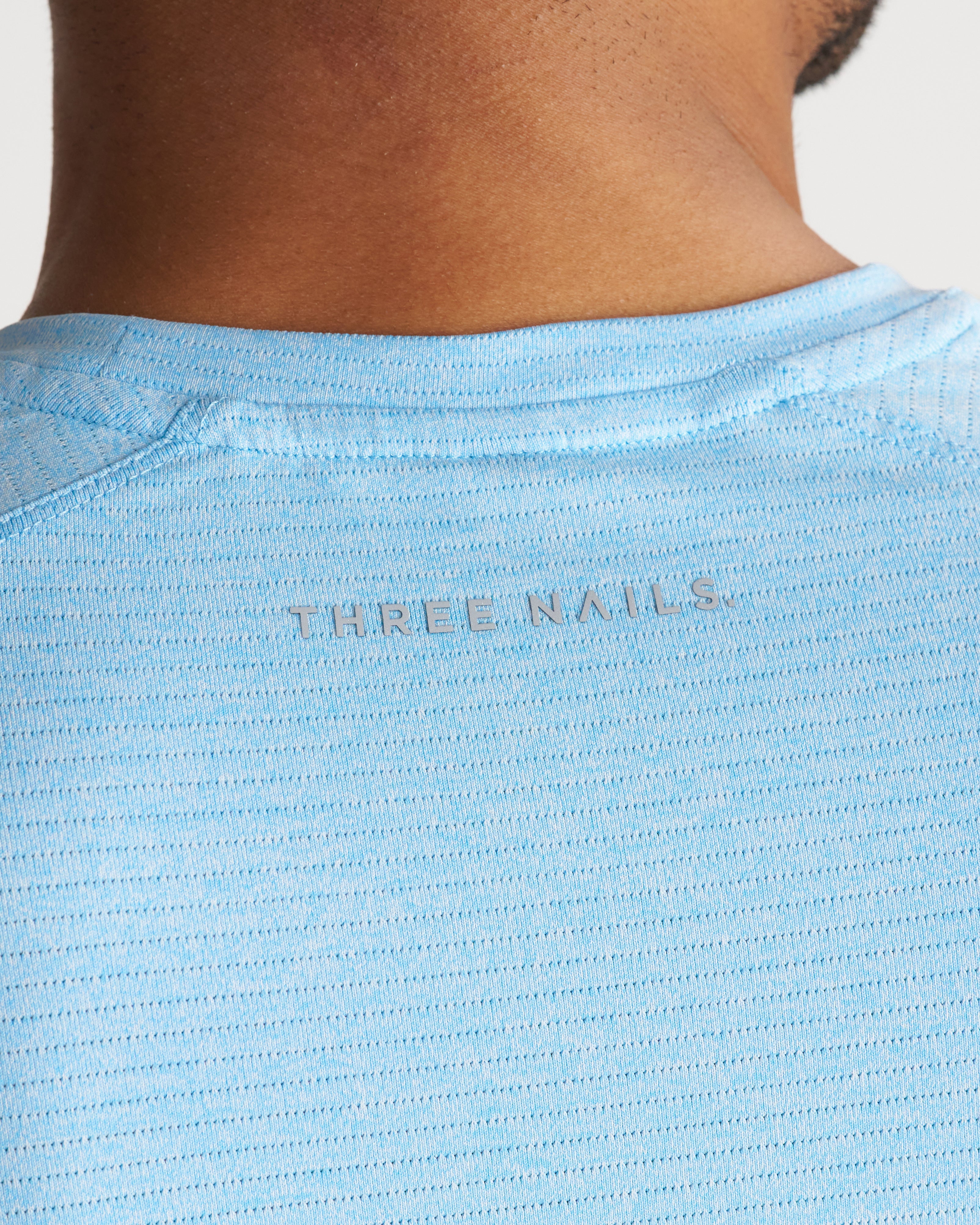Close-up back neck detail of the Aion-Lite Tee Cross in Sky Blue Marl showing the Three Nails logo label.
