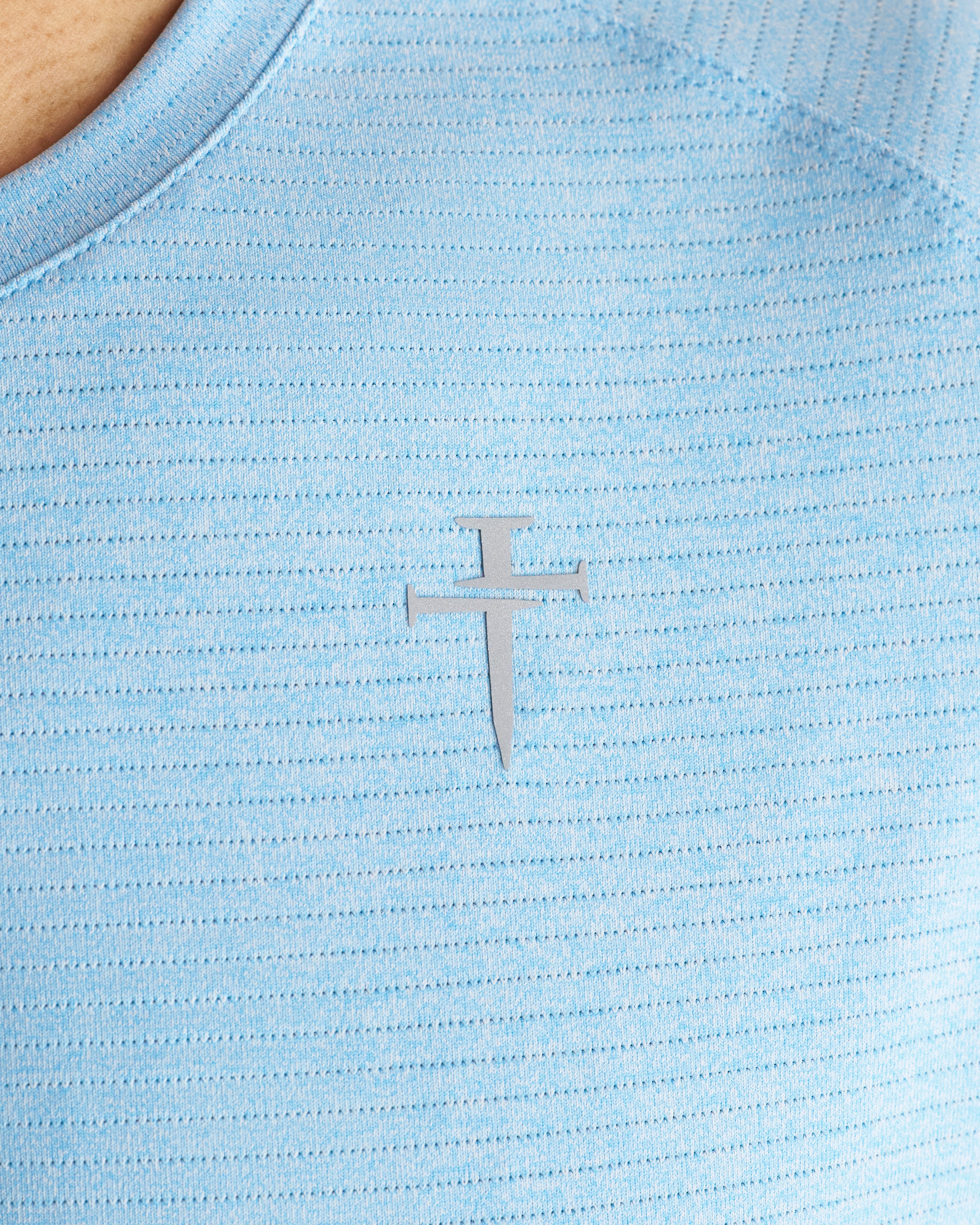 Close-up detail of the Aion-Lite Tee Cross in Sky Blue Marl showing the silver cross logo on textured fabric.