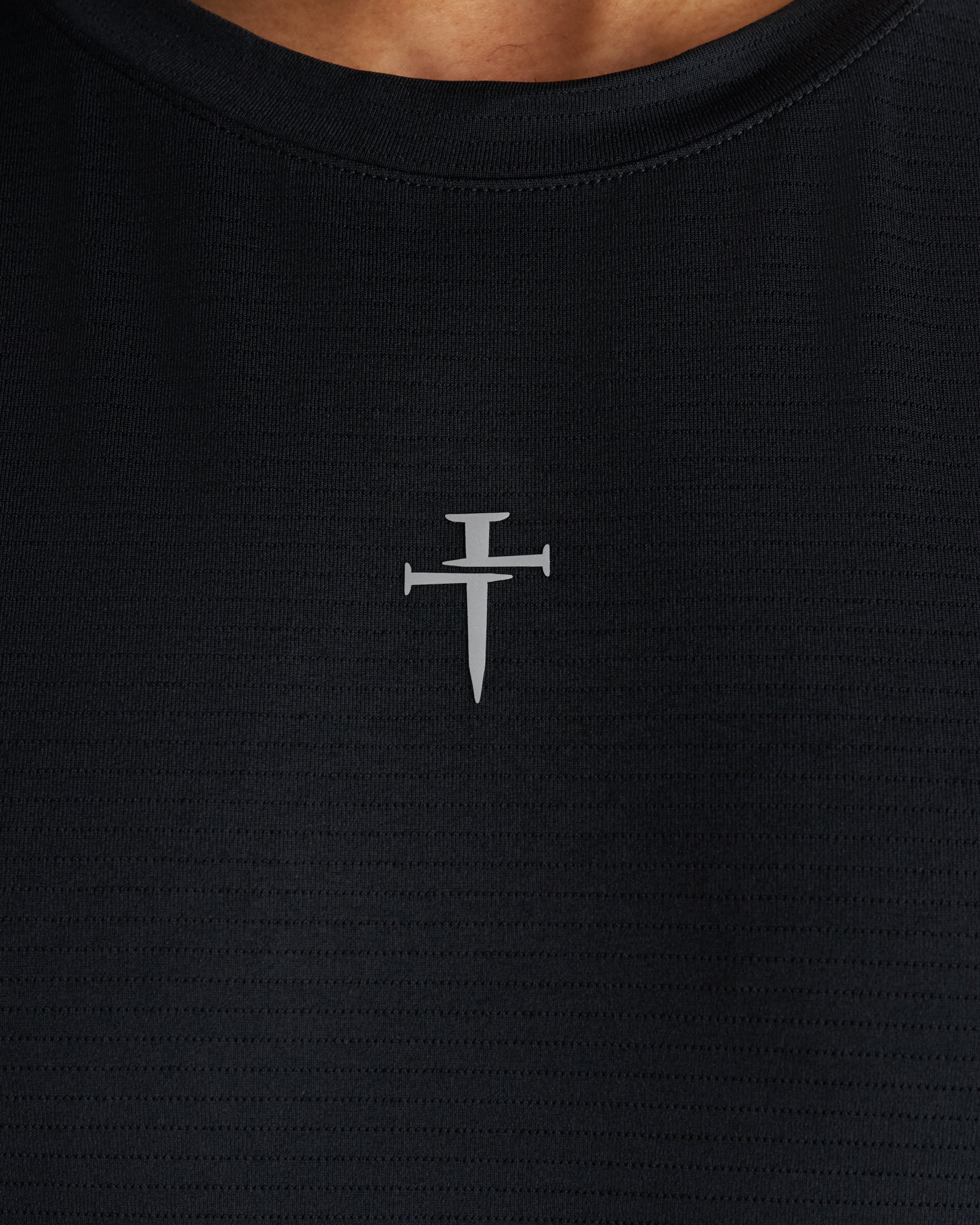 Close-up of the black Aion-Lite Tank chest area showing a small silver three nails cross logo on textured fabric.