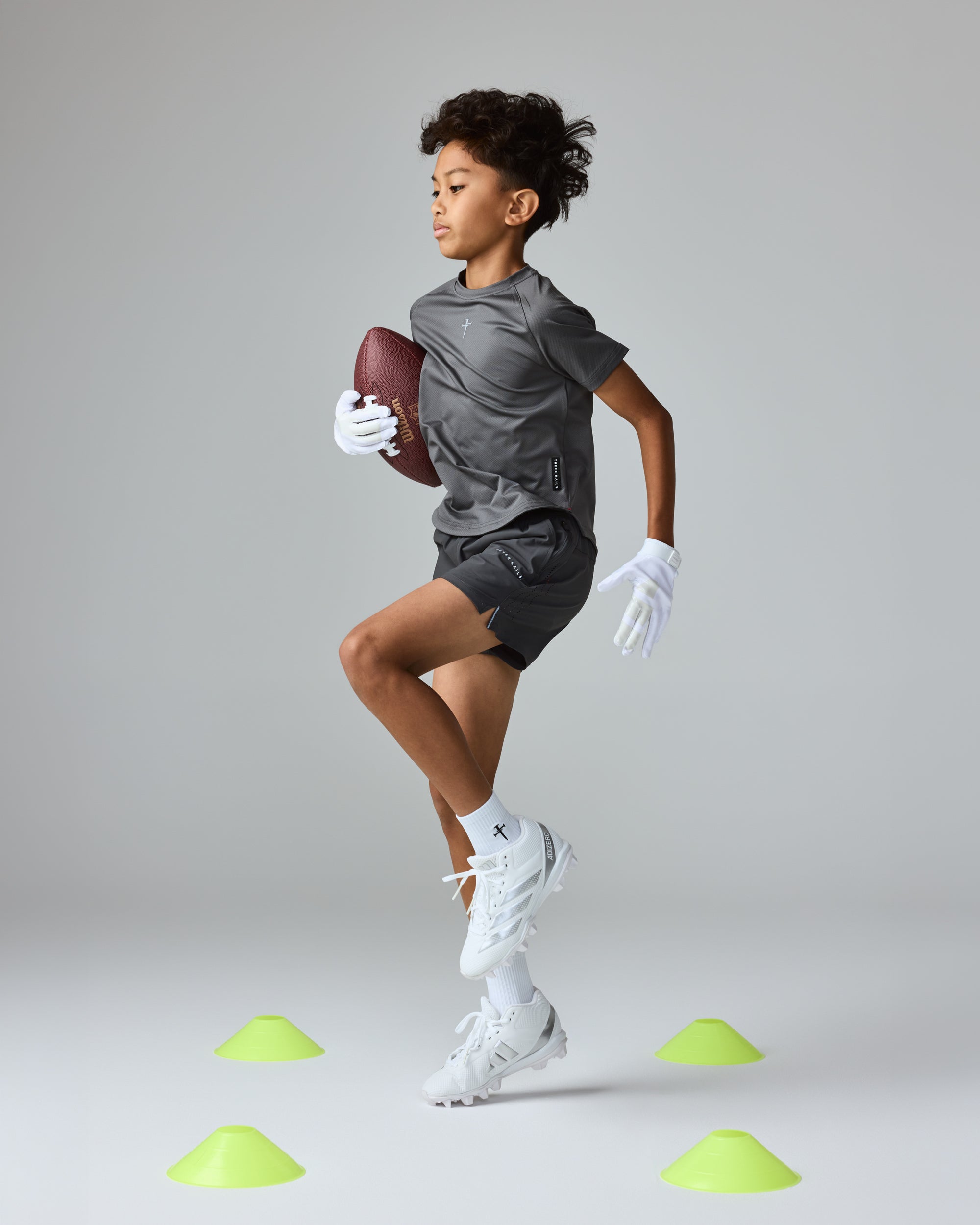 Youth Pro-Tech Tee Cross in charcoal on a young boy in a side-angle running pose holding a football.
