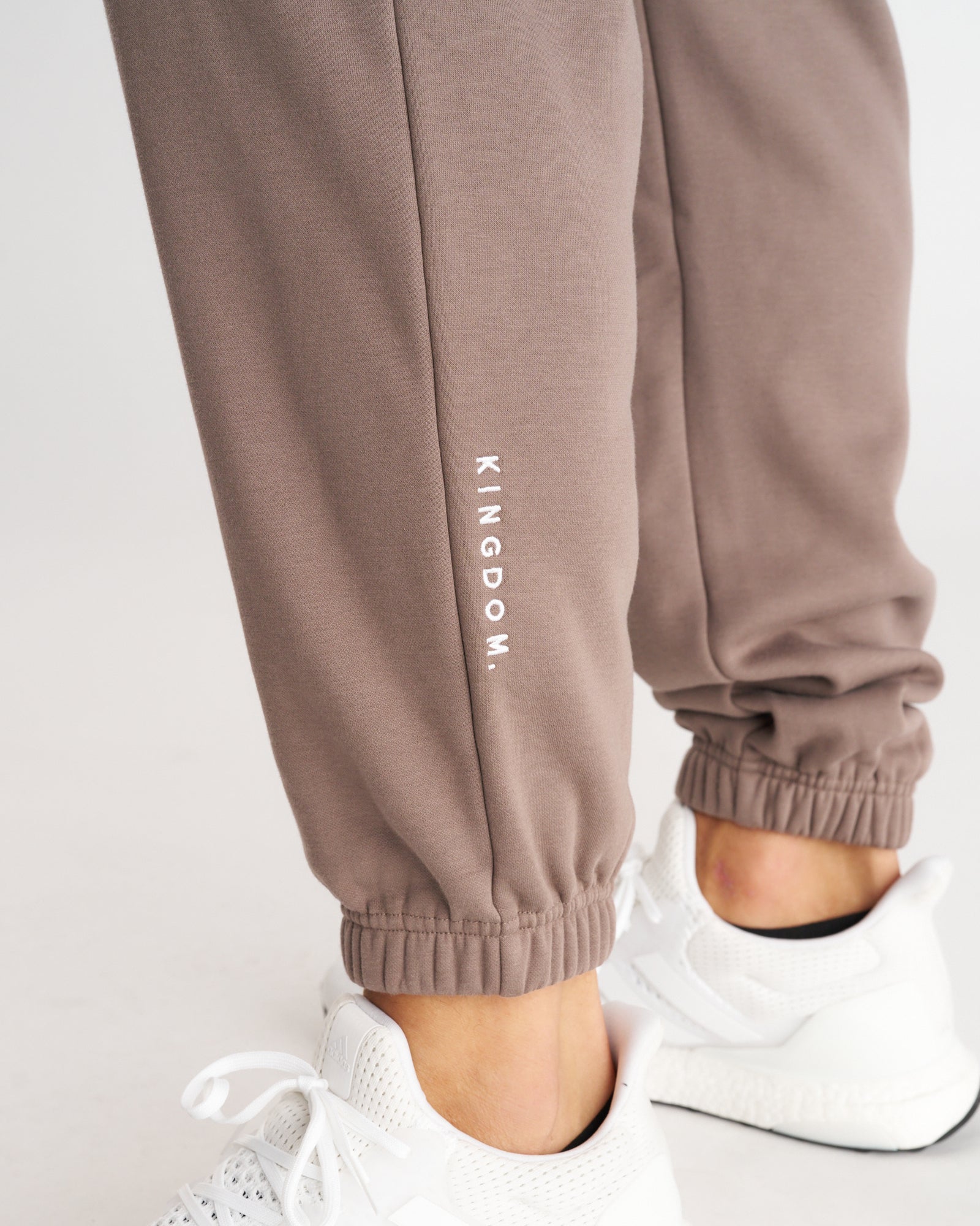 Close-up of Deep Taupe Signature Fleece Sweatpants showing ribbed ankle cuff and embroidered KINGDOM. text on lower leg.