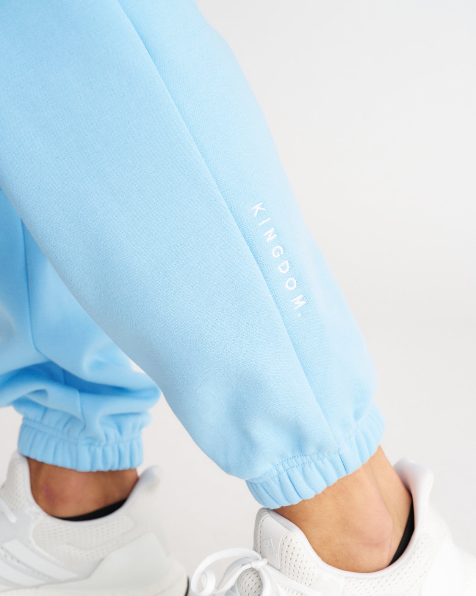 Close-up of Sky Blue Signature Fleece Sweatpants cuffed ankles with KINGDOM. logo, paired with white sneakers.