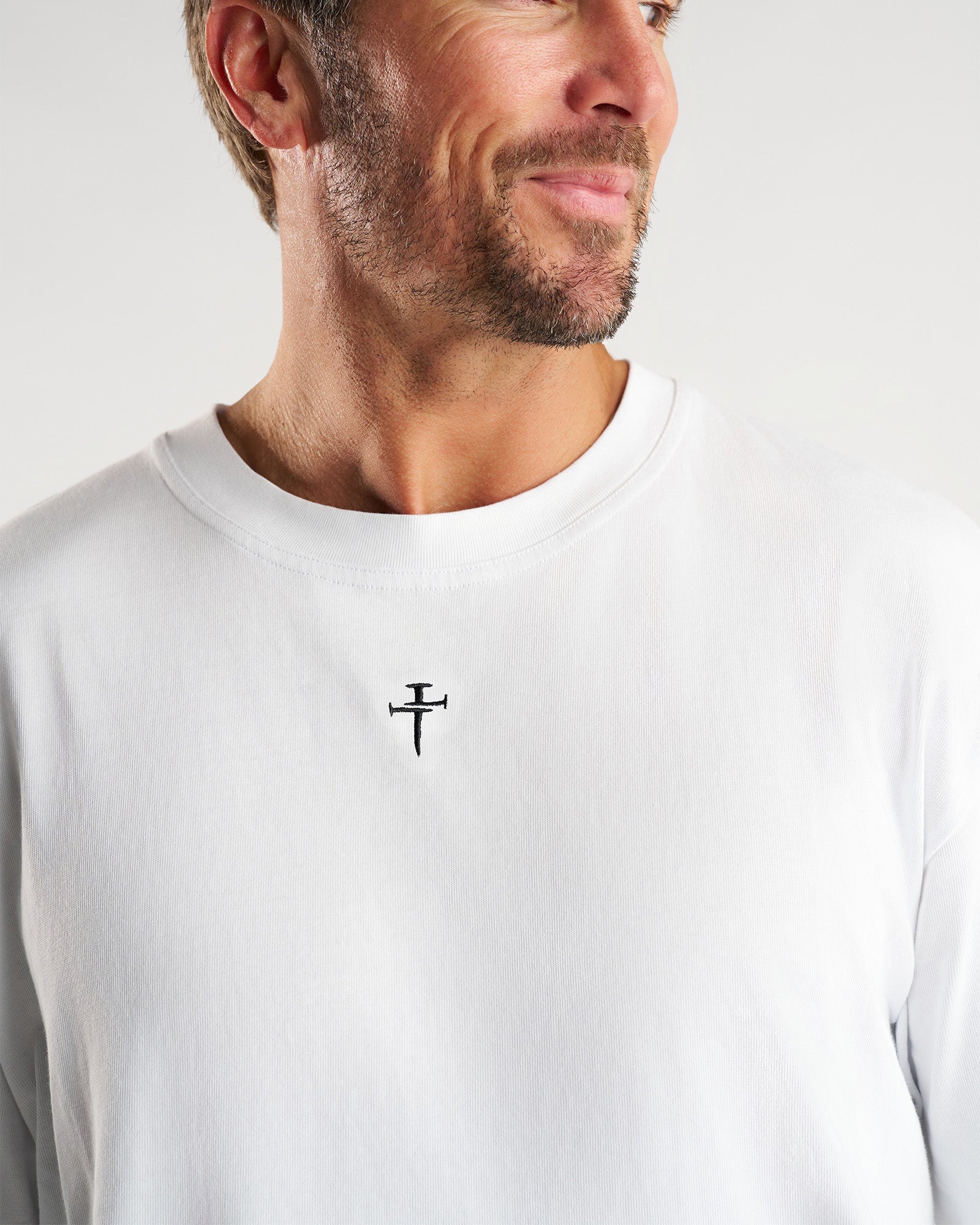 Close-up of white Heavyweight Cotton Oversized Tee FHG showing embroidered nail cross logo at chest center.