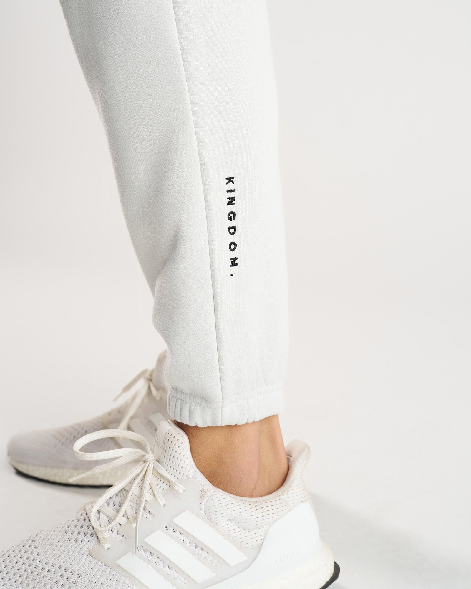 Close-up of Stone Signature Fleece Sweatpants ankle cuff with KINGDOM. text print, styled with white Adidas sneakers.