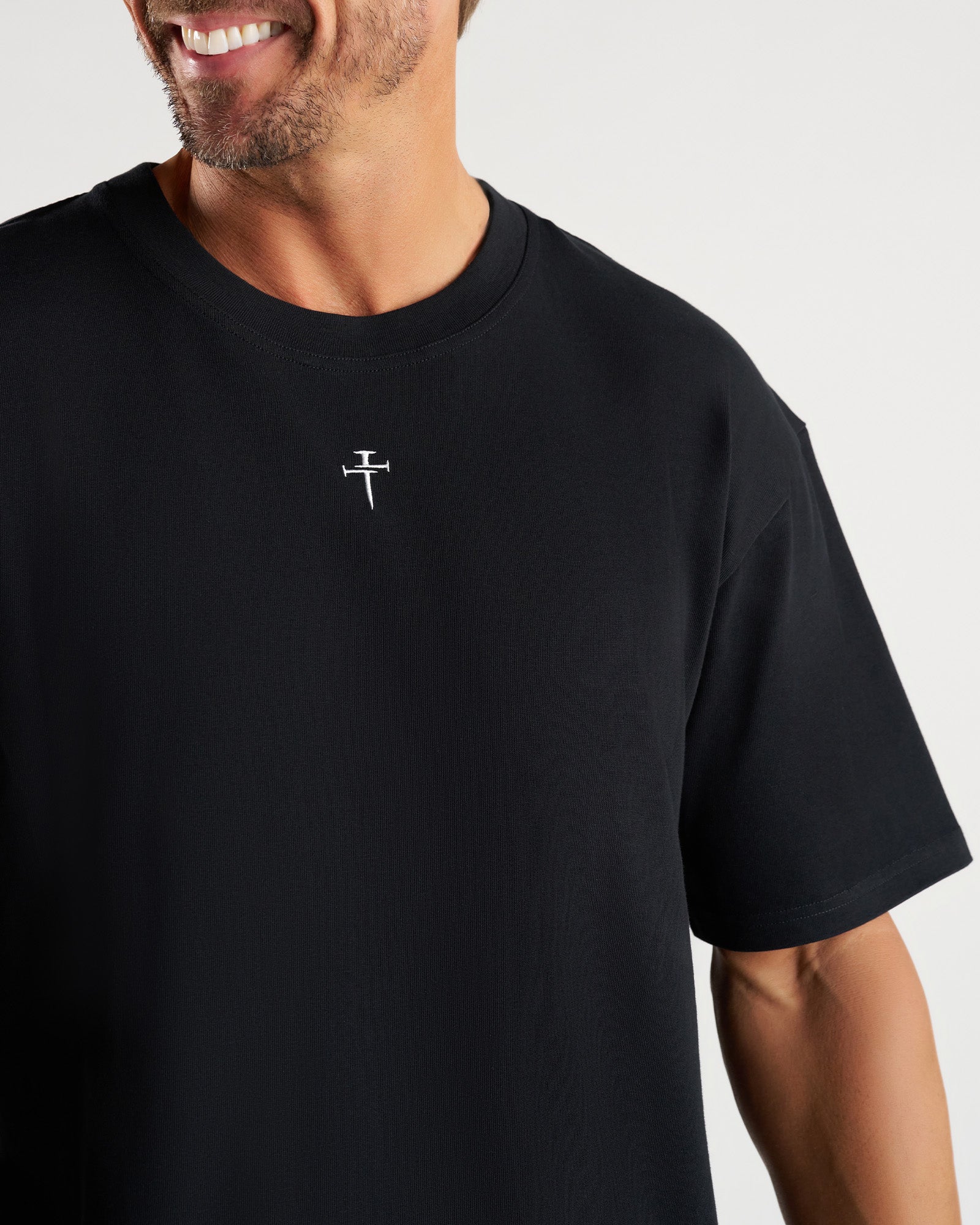 Close-up of black Heavyweight Cotton Oversized Tee showing embroidered nail cross logo on chest.