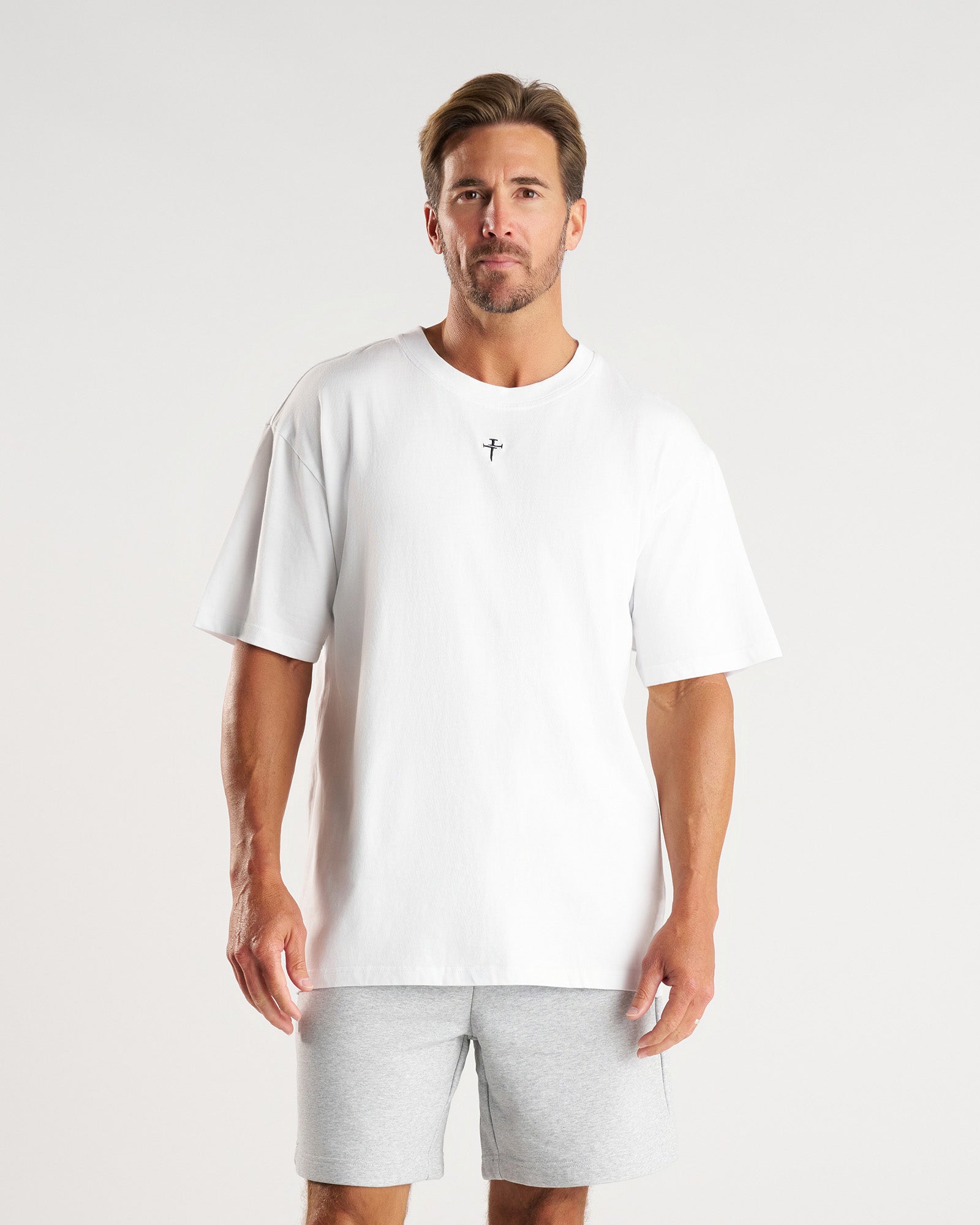 Man wearing white Heavyweight Cotton Oversized Tee FHG, front view, small cross logo at chest, paired with grey shorts.