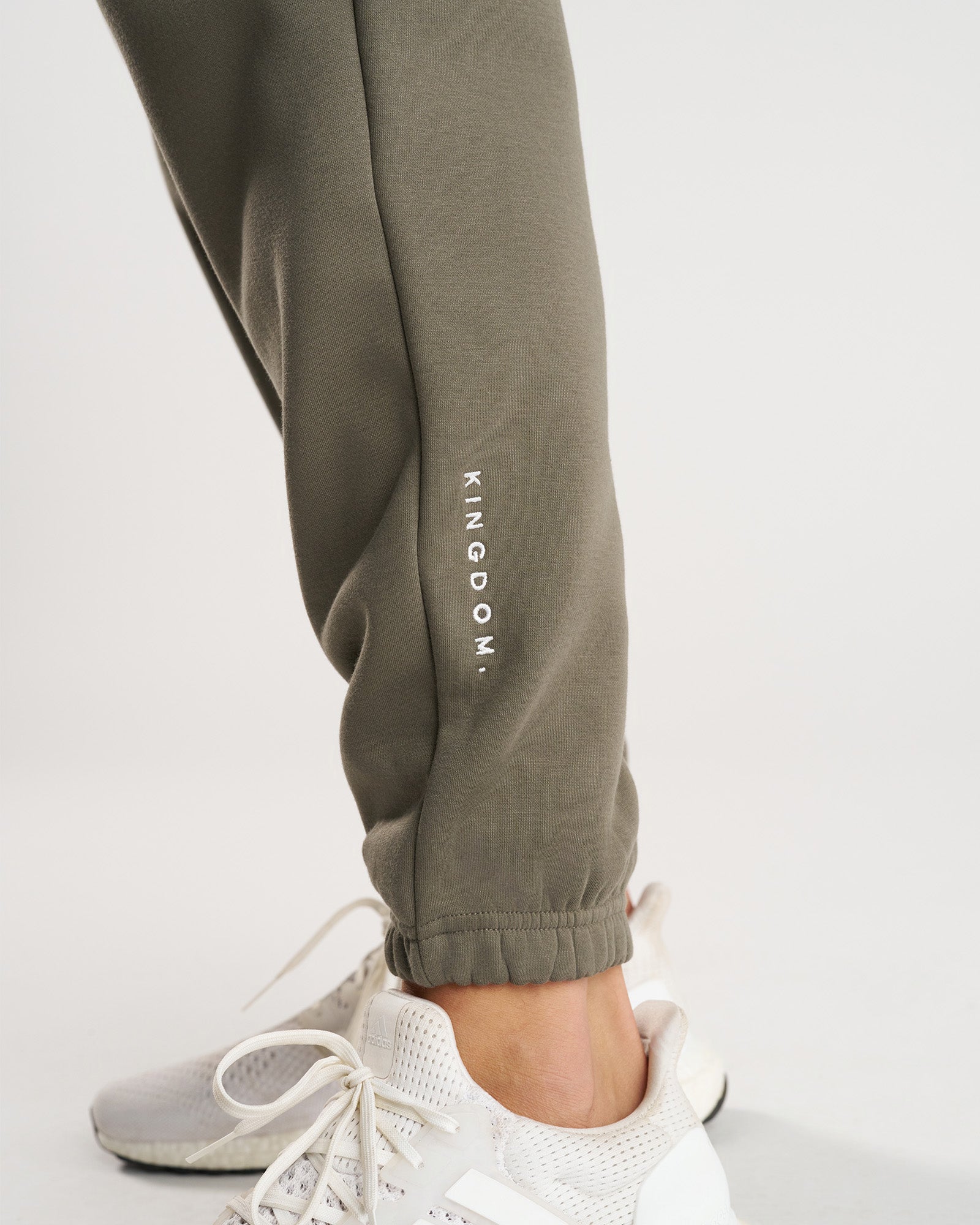 Close-up of Signature Fleece Sweatpants in Sage showing the ribbed cuff and embroidered KINGDOM. logo above white sneakers.