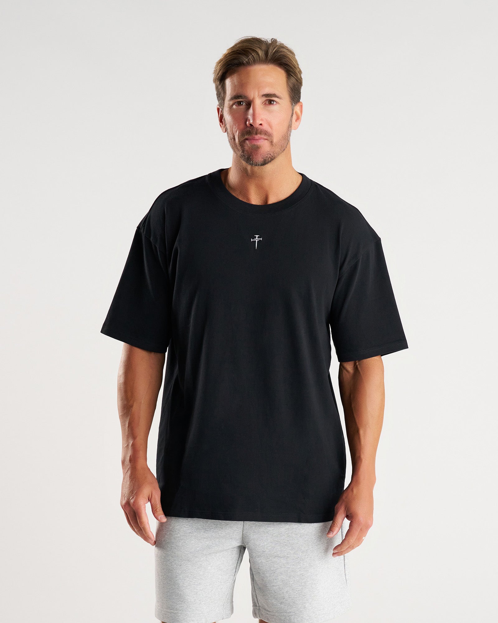 Man wearing black Heavyweight Cotton Oversized Tee, front view, small cross logo at chest, paired with grey shorts.
