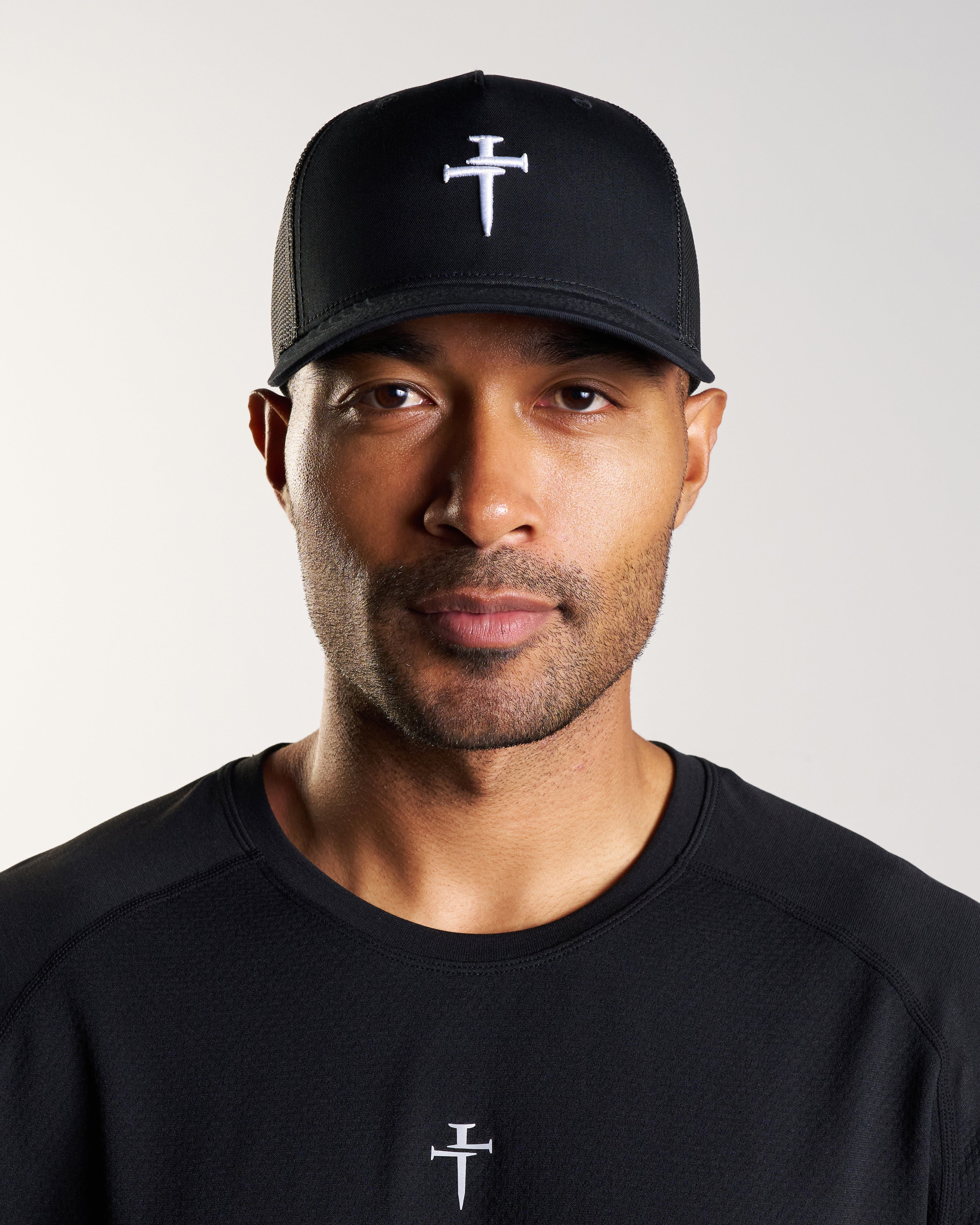 Man wearing black Signature Trucker Hat with white nail cross logo, front view, paired with matching black tee.