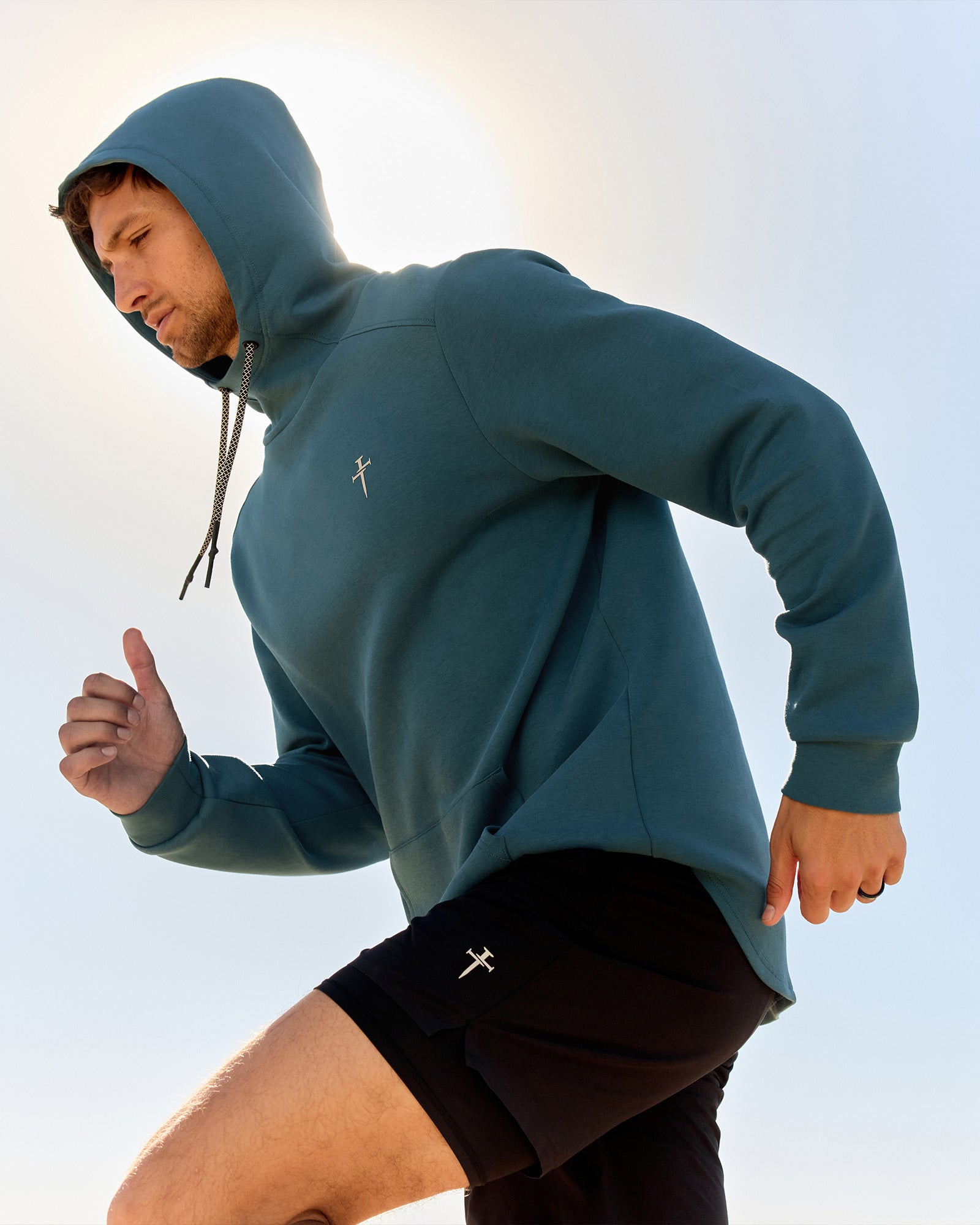 Man running in the Pro-Tech Hoodie in Teal, side angle, paired with black shorts against a bright sky.