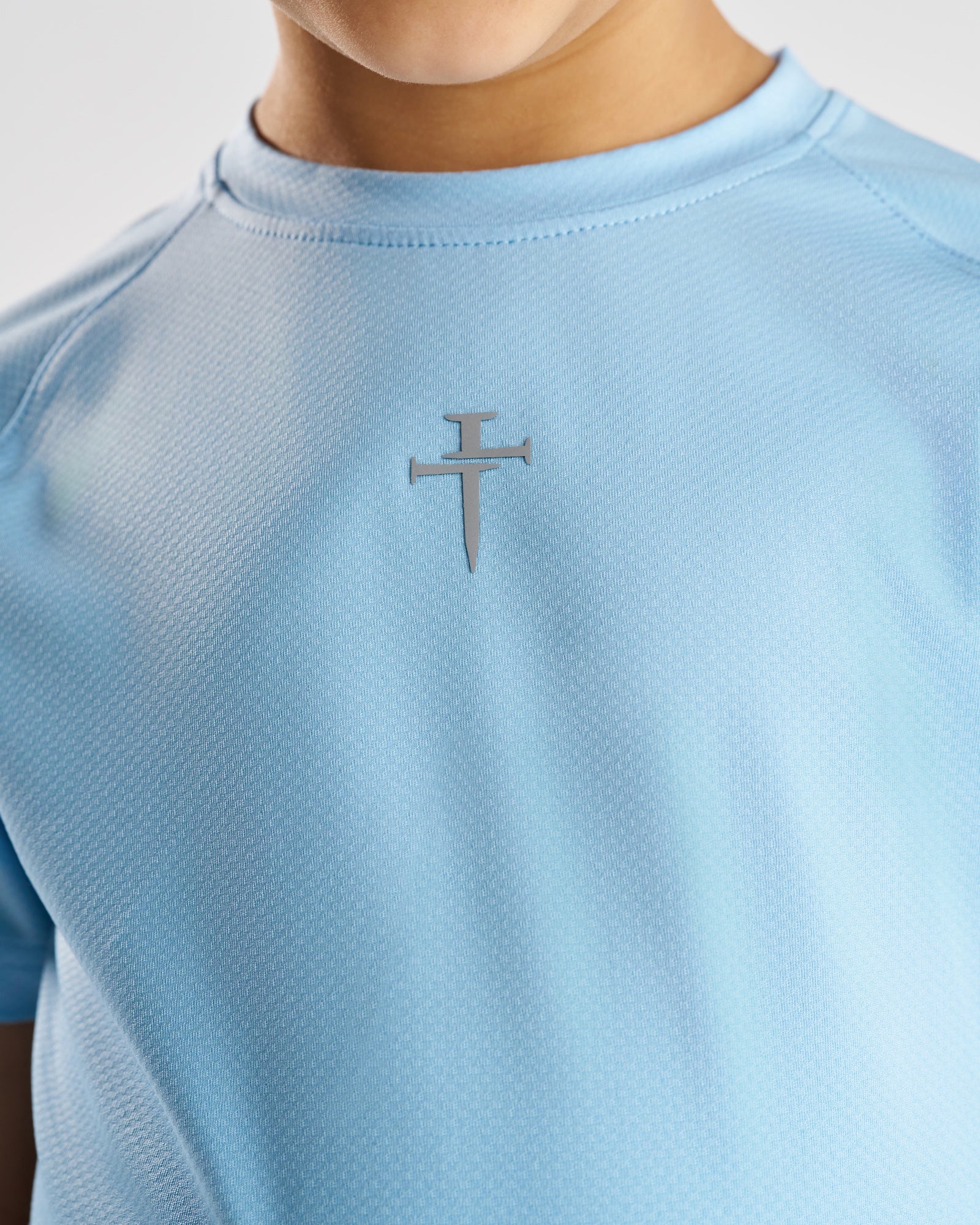 Close-up of Sky Blue Youth Pro-Tech Tee Cross showing the silver nail cross logo on chest.