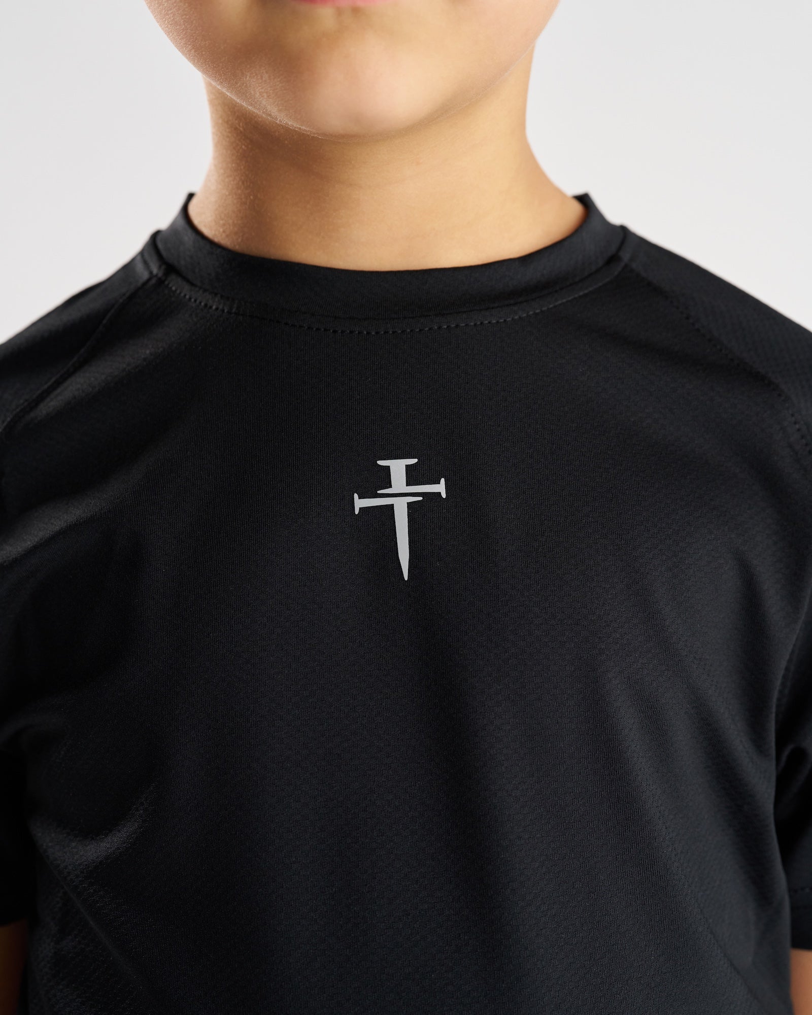 Close-up of the Youth Pro-Tech Tee Cross in black showing the silver nail cross logo on chest.