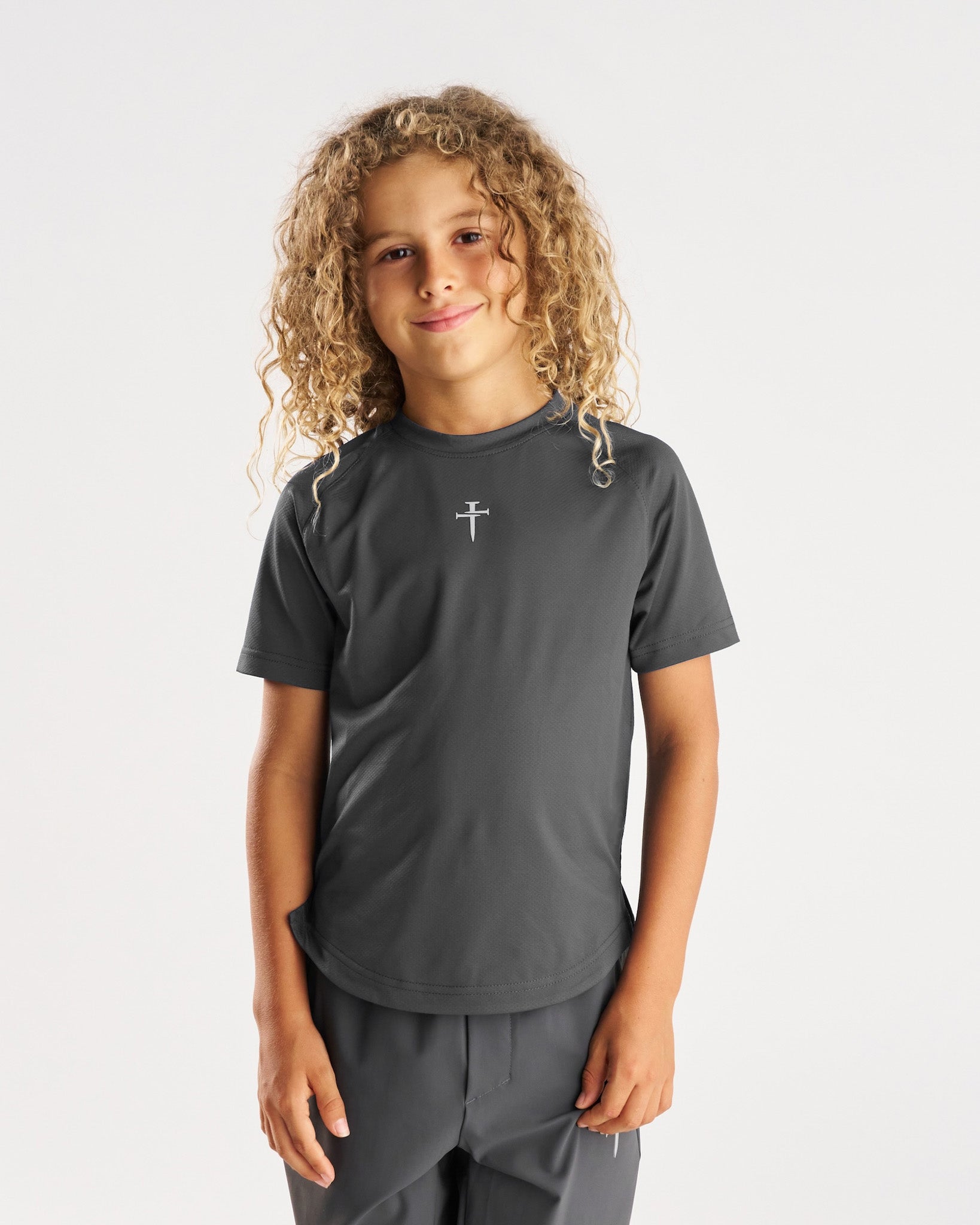 Youth Pro-Tech Tee Cross in Space Grey, front view on young model with curly hair, small cross logo at chest.
