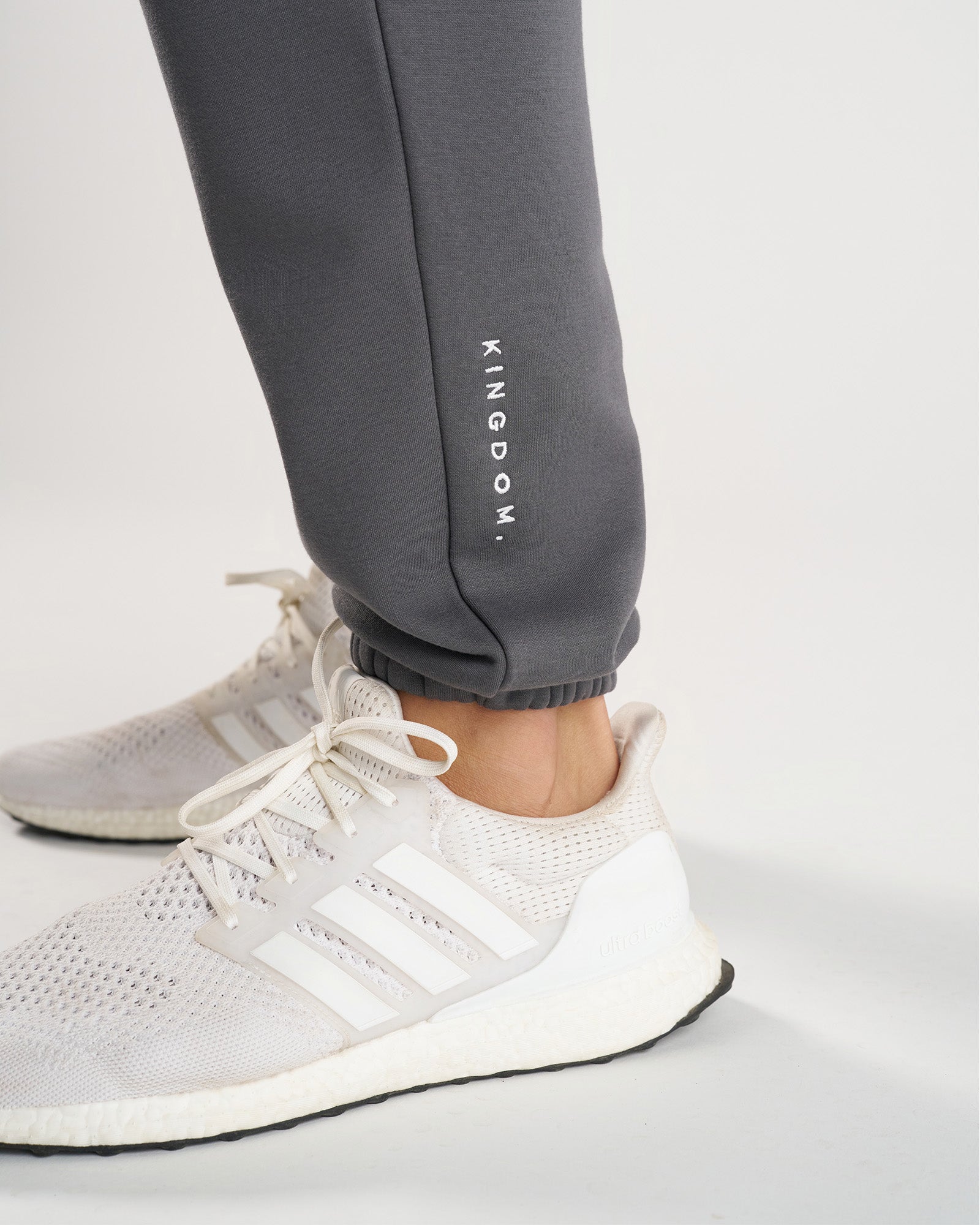 Close-up of Space Grey Signature Fleece Sweatpants cuffed ankle with KINGDOM. text, paired with white Adidas sneakers.