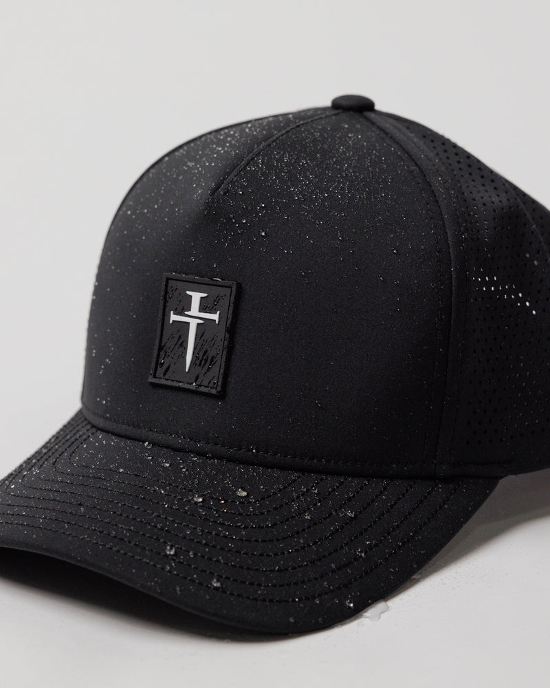 Close-up of a black Perforated Hat Bundle Pack showing a rubber cross logo patch and speckled water-resistant fabric.