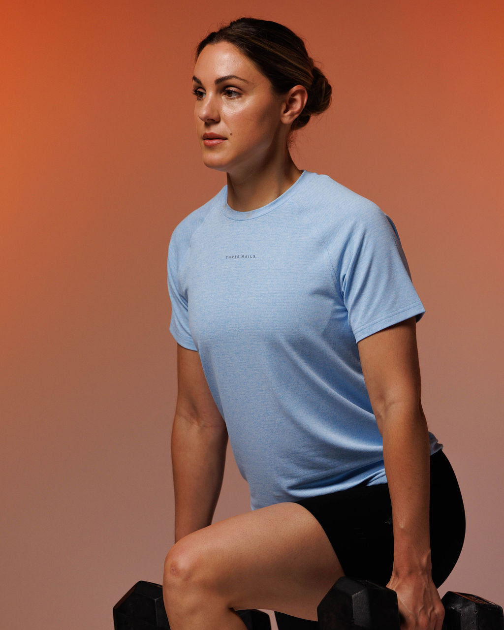 Women's Aion-Lite Tee in Sky Blue Marl, model seated at side angle holding dumbbells, black shorts.