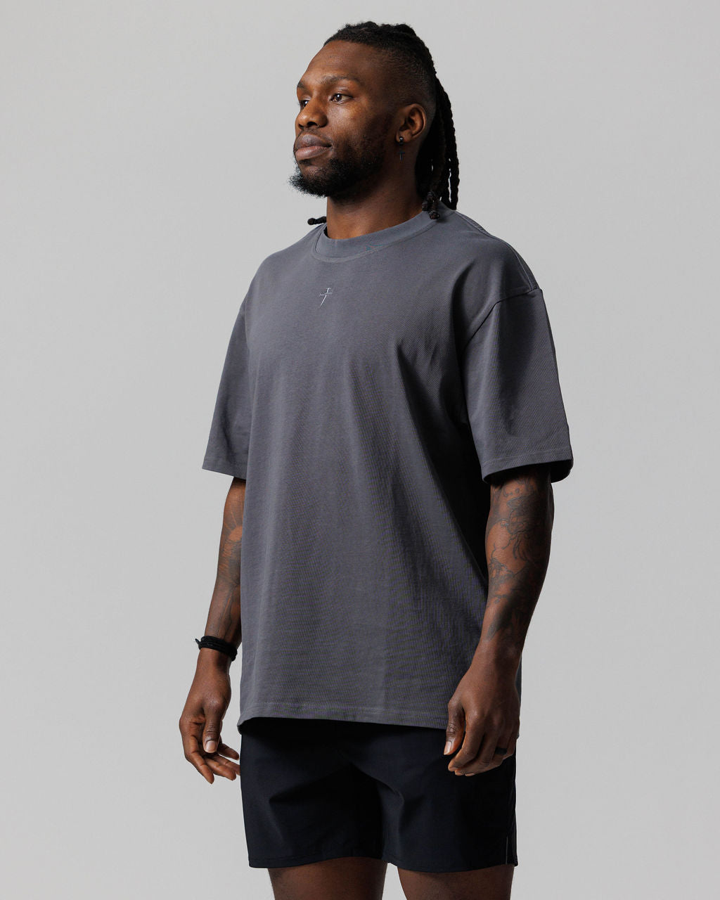 Man wearing Space Grey Heavyweight Cotton Oversized Tee with small cross logo, three-quarter front view, paired with black shorts.