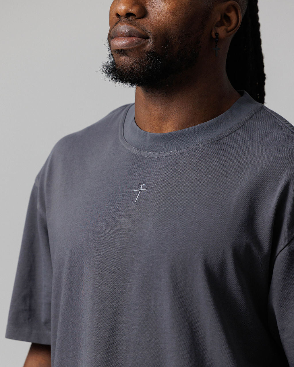 Close-up of Space Grey Cross Heavyweight Oversized Tee showing embroidered nail cross logo at chest on male model.