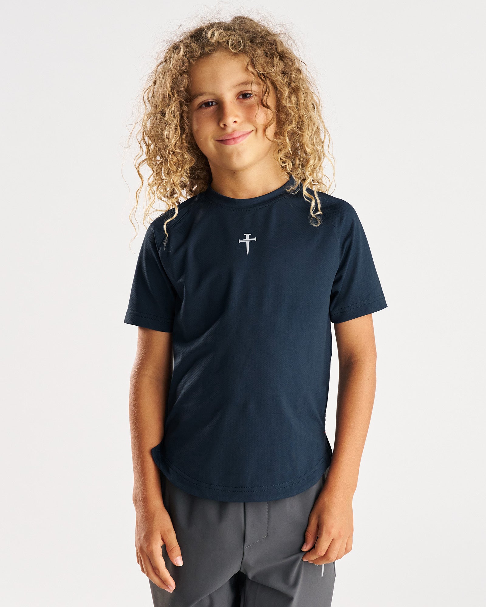 Youth Pro-Tech Tee Cross in Midnight Navy, front view on a young model with curly hair, small cross logo at center chest.