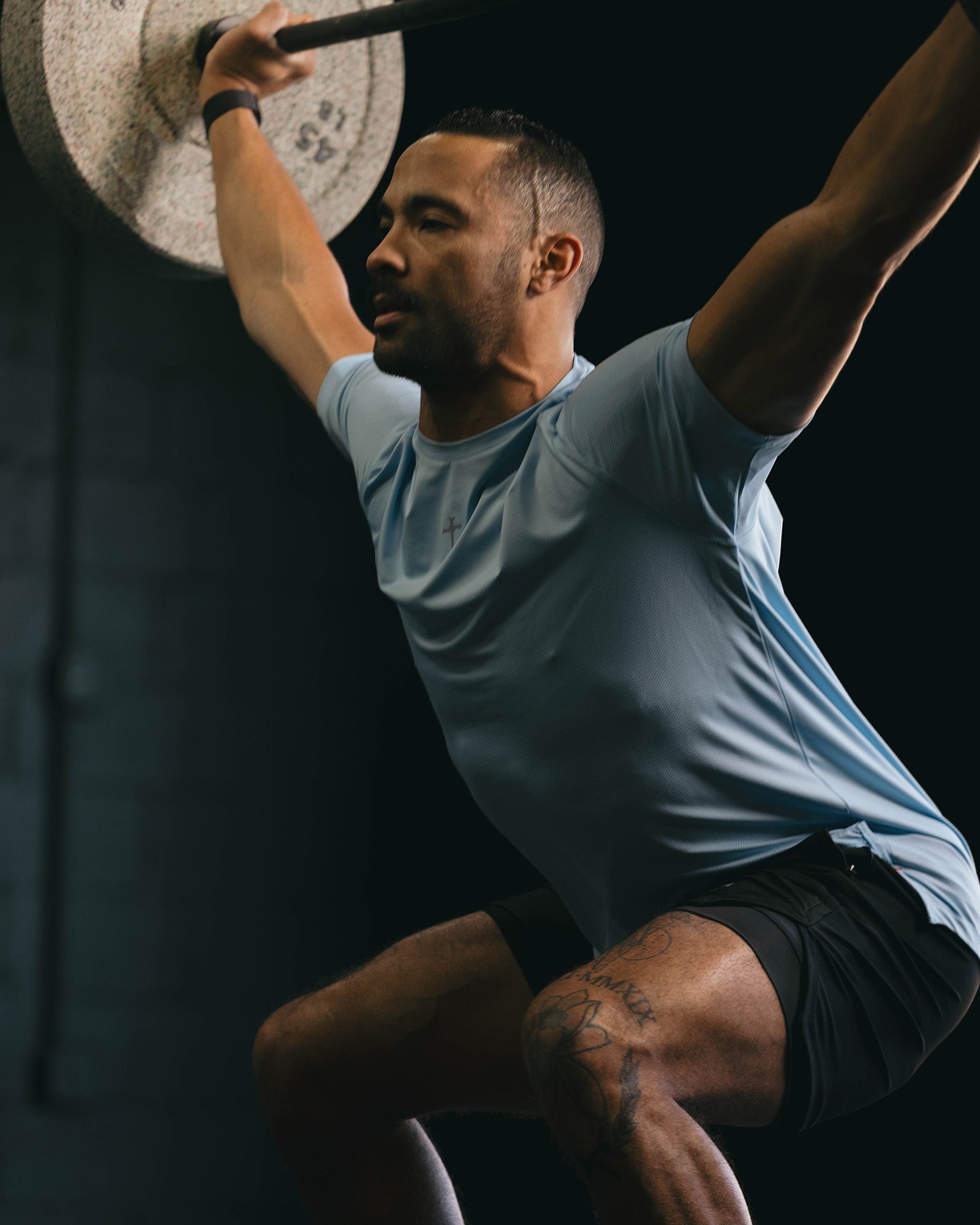 Male athlete performing overhead lift wearing Pro-Tech Tee Cross in sky blue, front angle view with tattoos visible.