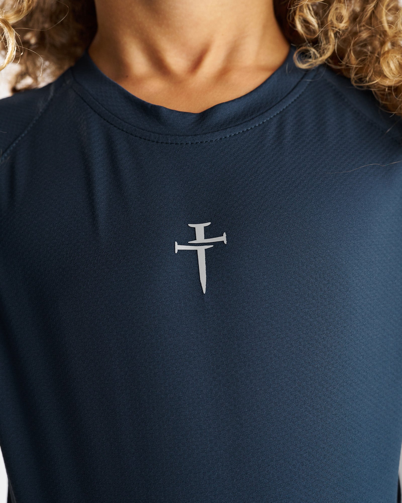 Close-up of the silver nail cross logo on the Youth Pro-Tech Tee in Midnight Navy.