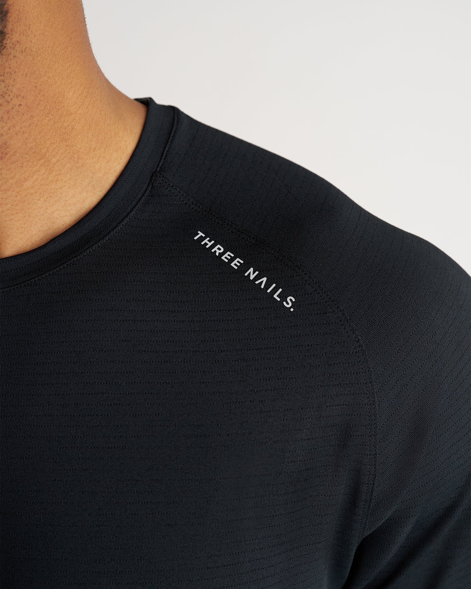 Close-up detail shot of the Aion-Lite Tee in black showing the Three Nails chest logo and textured fabric.