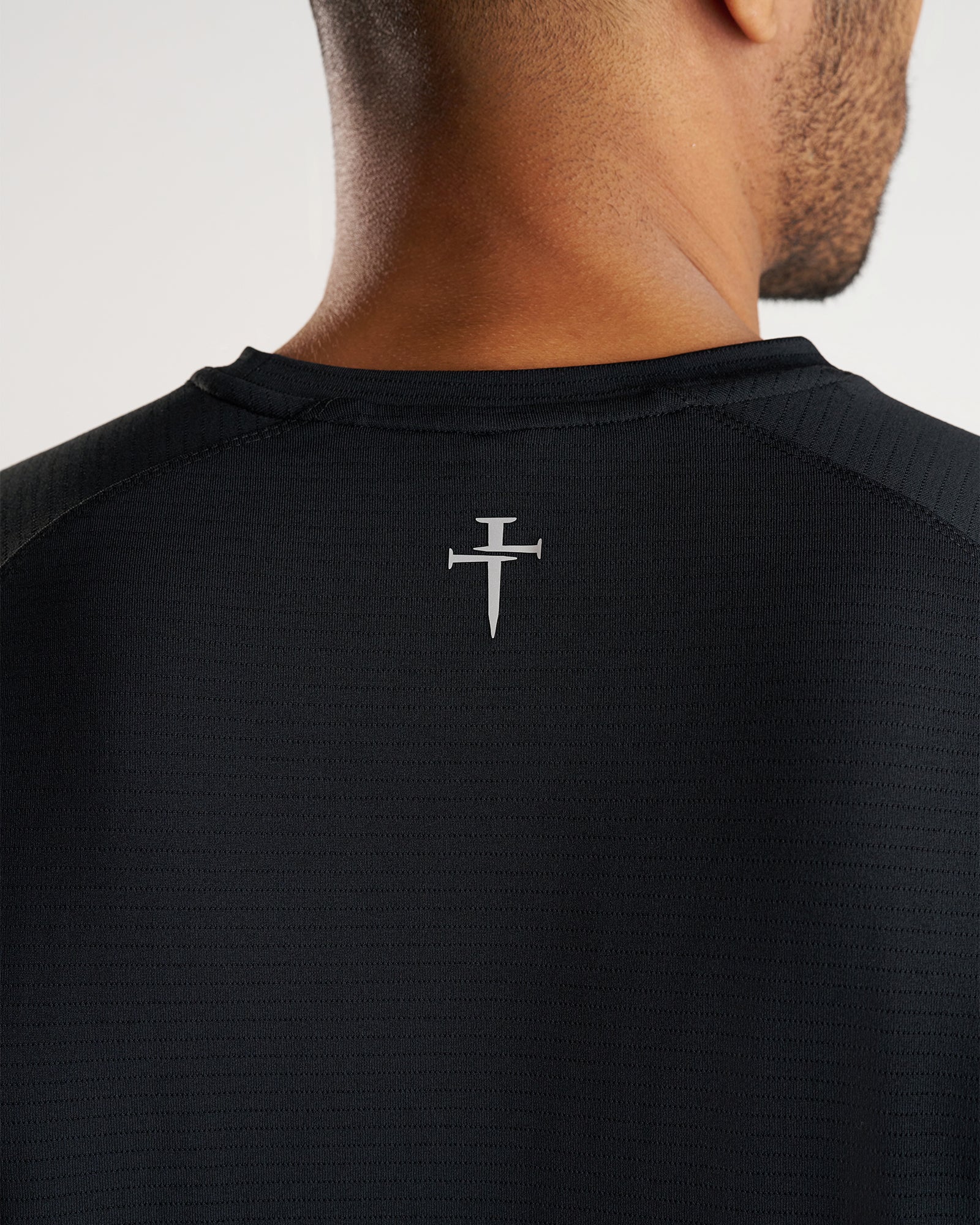 Close-up back view of the Aion-Lite Tee Three Nails in black showing the small silver nail cross logo below the collar.