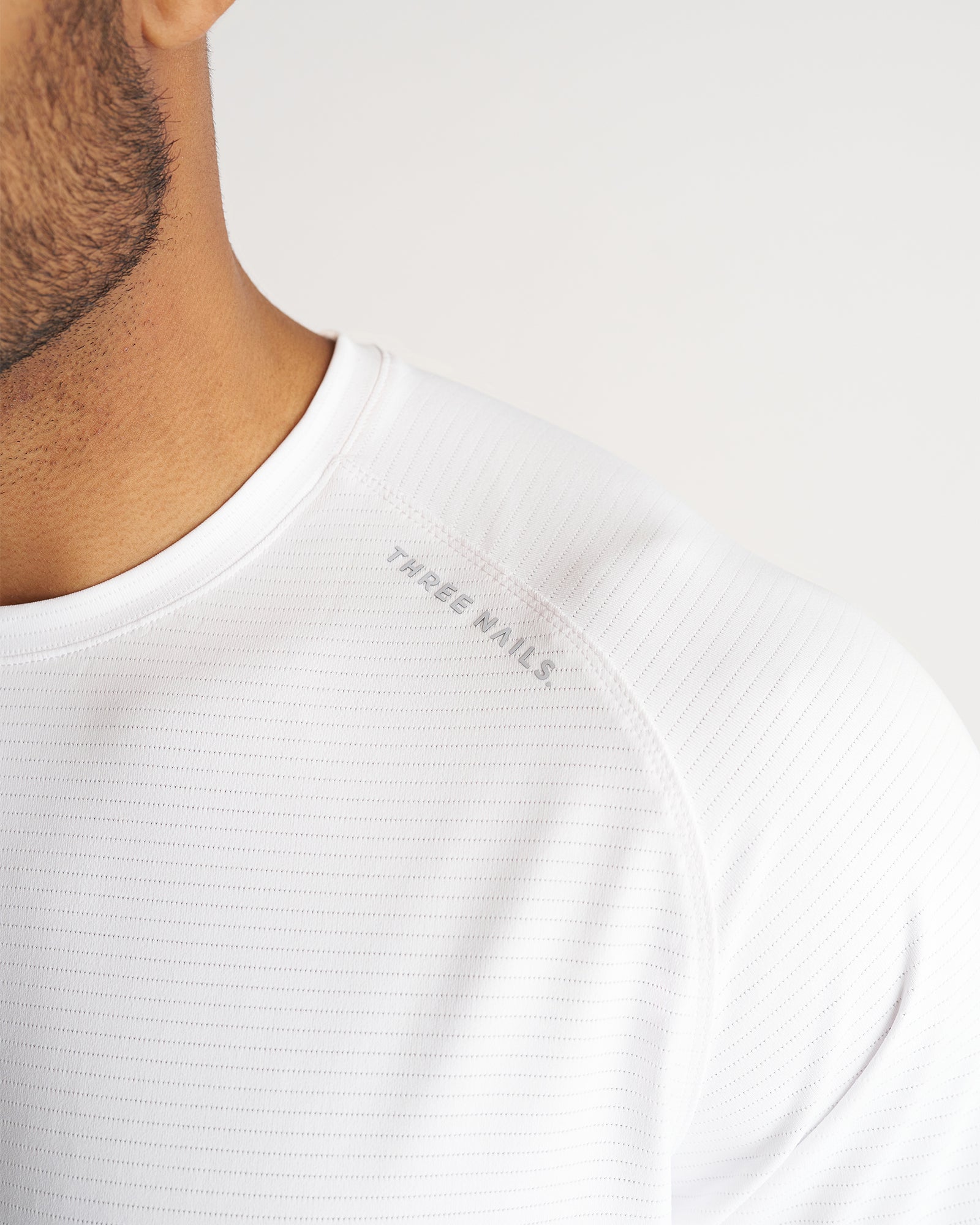 Close-up detail shot of the Aion-Lite Tee in white showing ribbed texture, raglan seam, and Three Nails logo at the shoulder.