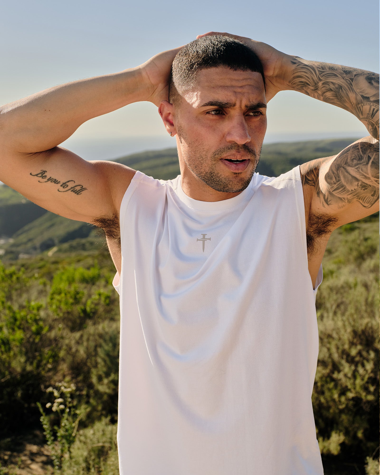 Male model wearing white Core Raglan Cutoff tank top, front view, arms raised behind head, outdoor hillside setting.