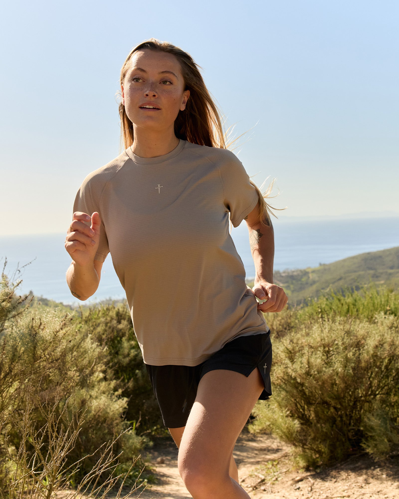 Woman running outdoors in the Aion-Lite Tee Cross in Sand with black shorts, front view.