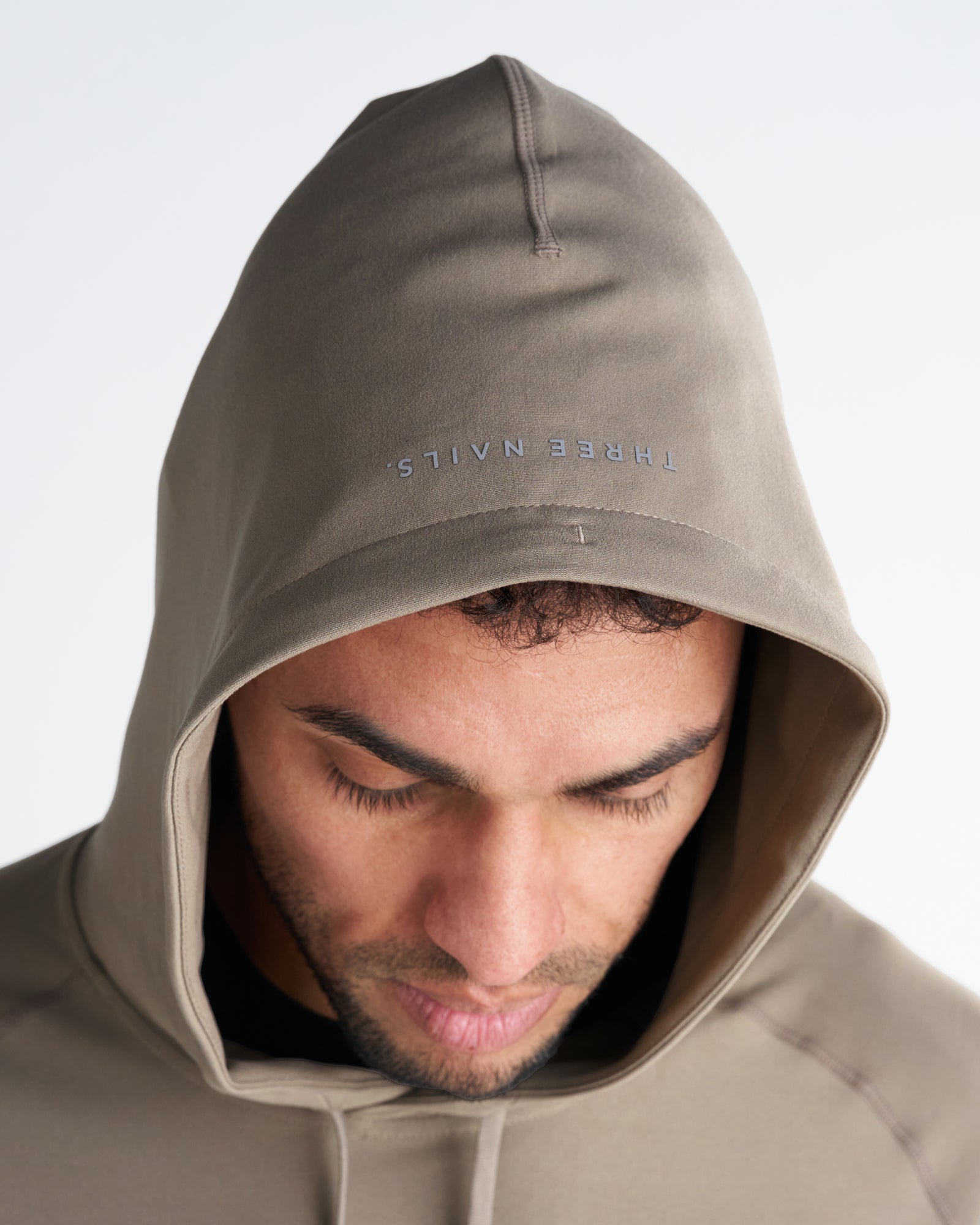 Close-up of Haven Performance Hoodie in Sand, showing hood detail with mirrored Three Nails logo on brim, model looking down.