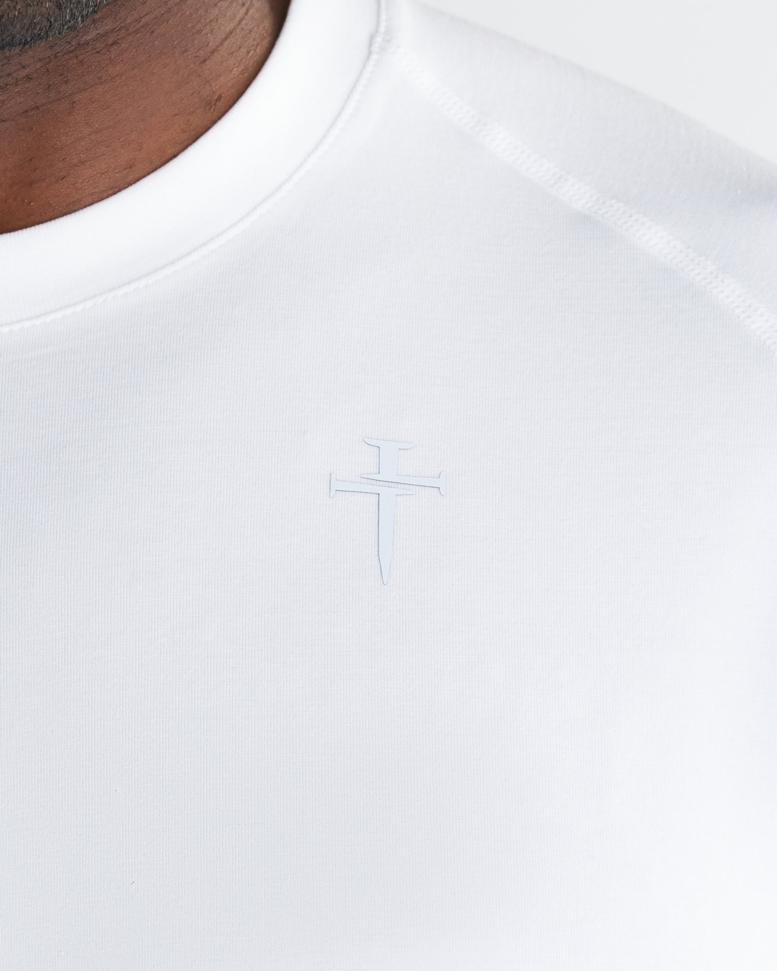 Close-up of the white Core Tee chest area showing a subtle tonal Three Nails cross logo on white fabric.