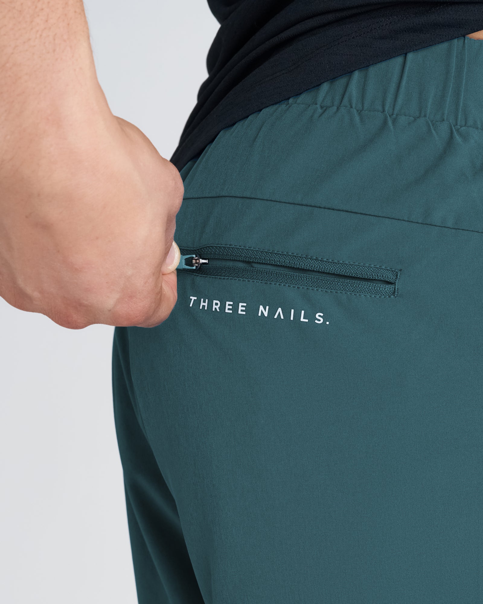 Close-up of Pulse Tech Linerless Short in teal showing zippered back pocket with Three Nails branding.