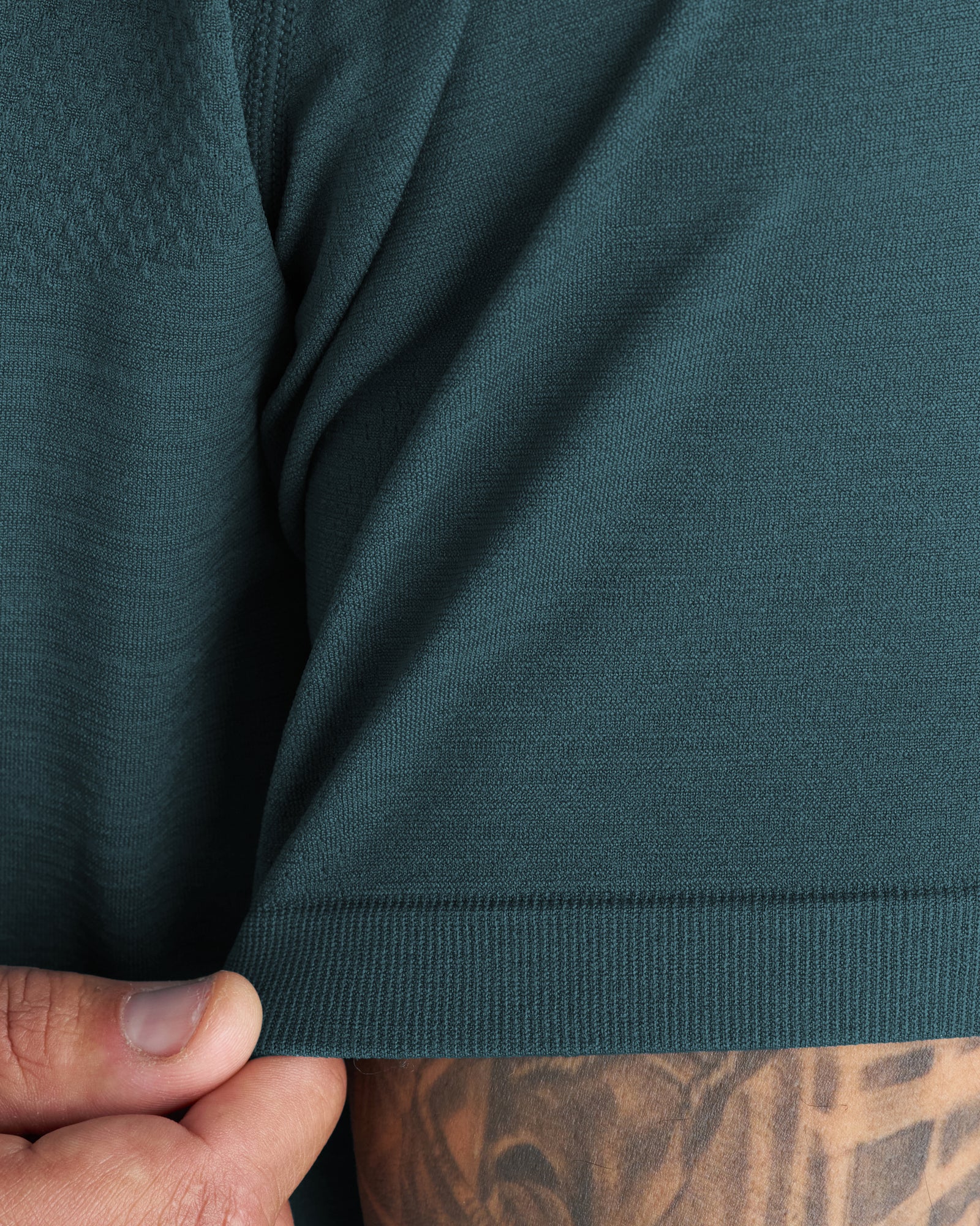Close-up detail of the Seamless Tee in Teal showing ribbed hem and fine knit texture with a tattooed hand visible.
