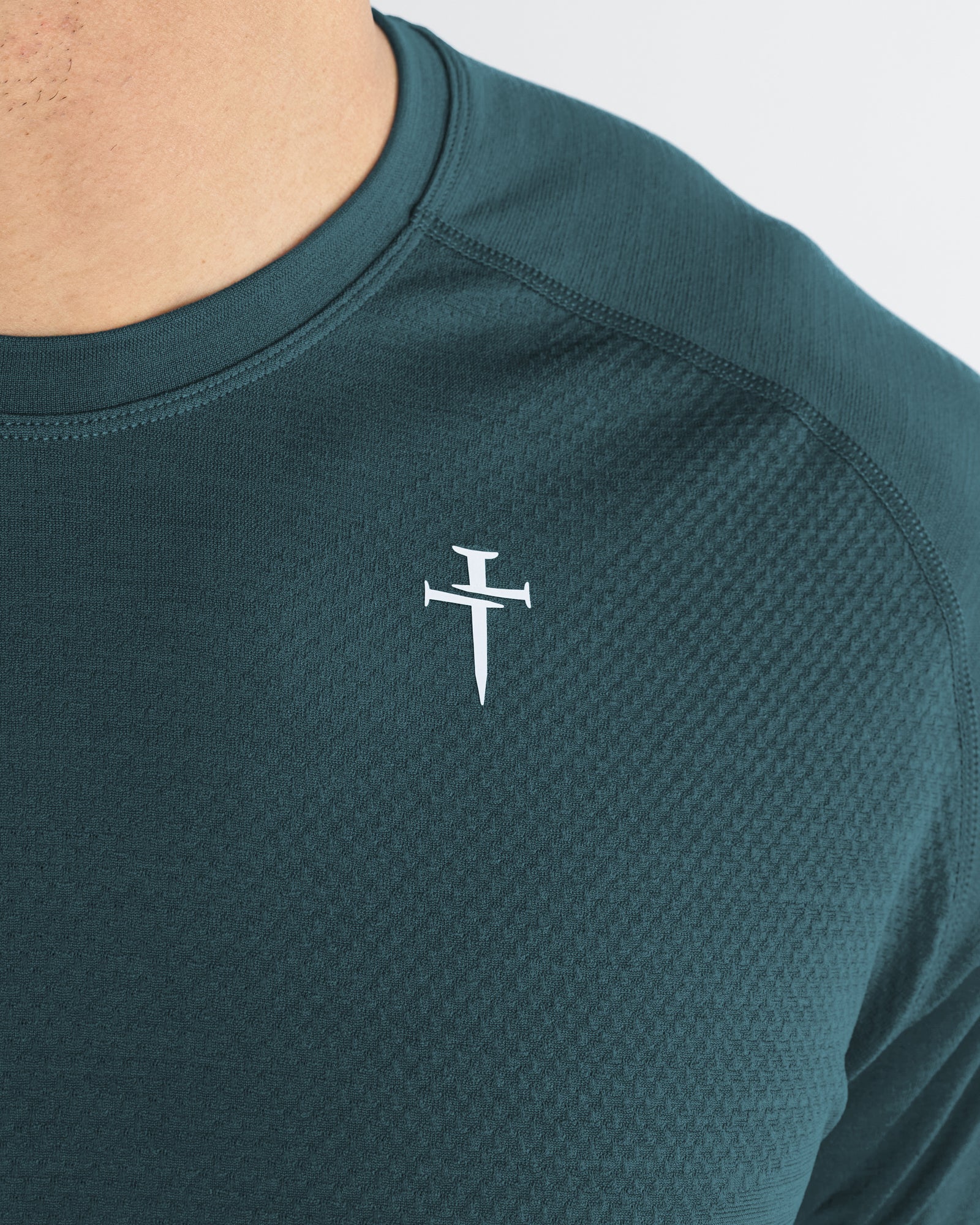 Close-up detail of the Seamless Tee in Teal showing the three nails cross logo and textured shoulder panel.