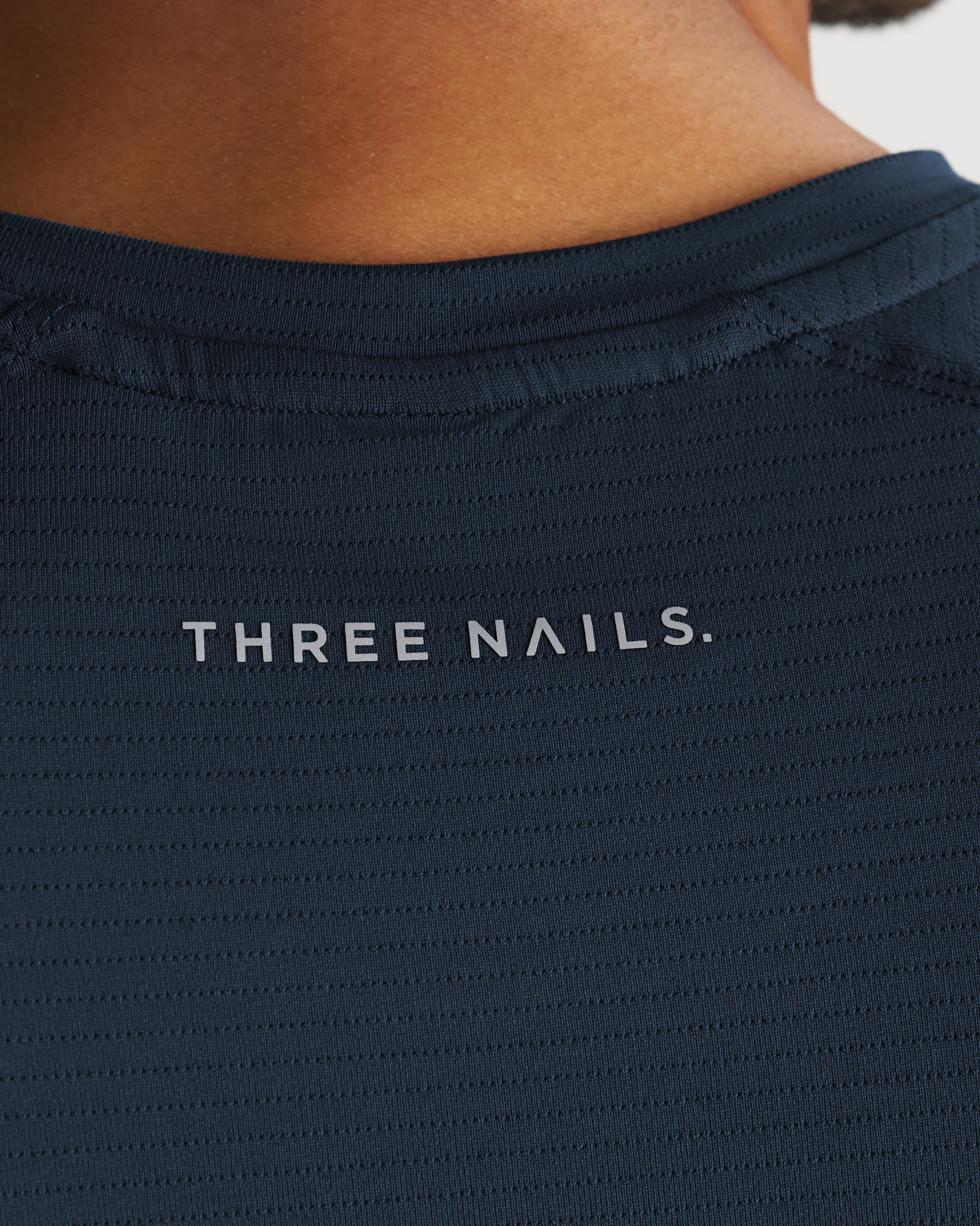 Close-up of the back collar area of the Aion-Lite Tee Cross in Midnight Navy showing the Three Nails. logo detail.